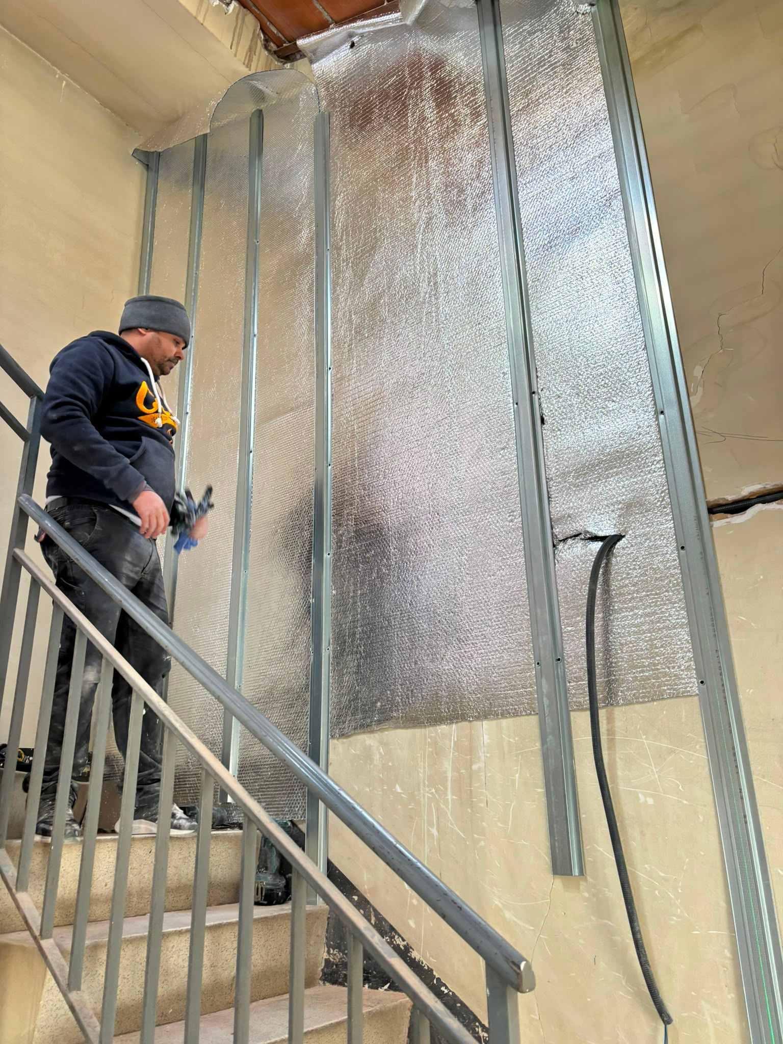 Trabajador en una escalera junto a paneles de pared cubiertos de aislamiento plateado y cables colgantes.