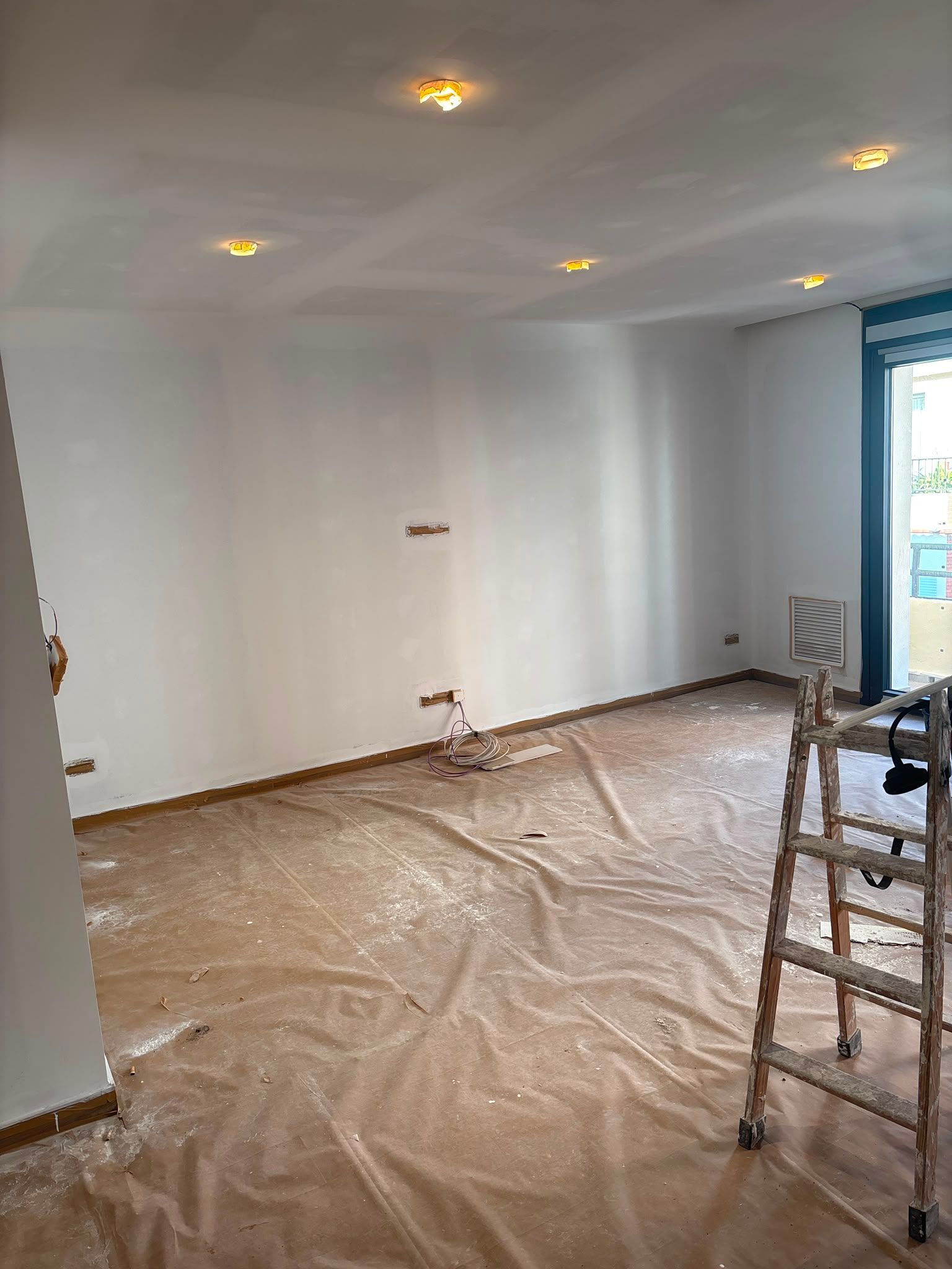 Habitación vacía en obras, con el suelo cubierto de plástico y una escalera cerca de la ventana.