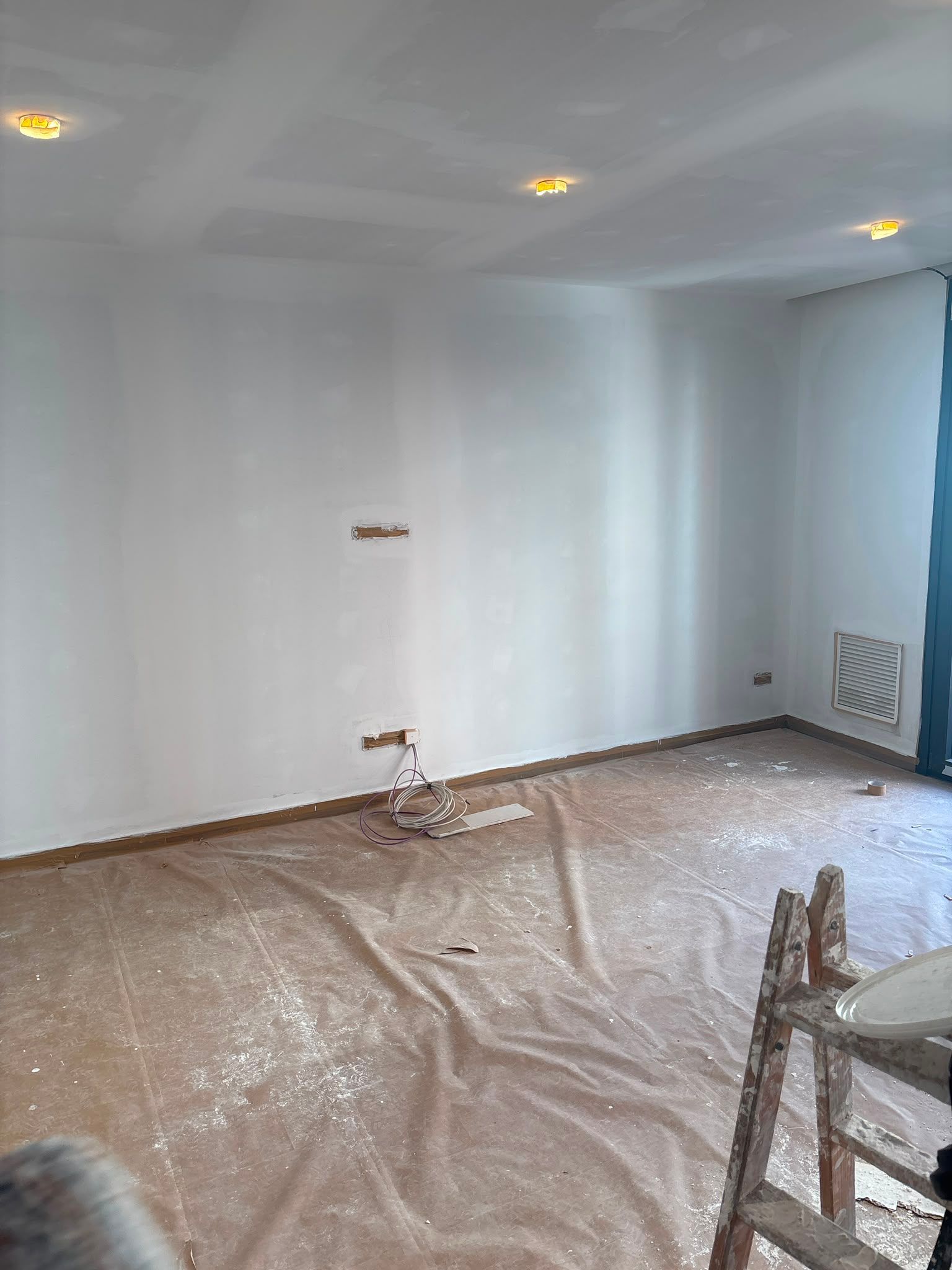 Habitación blanca vacía en proceso de renovación, con suelo cubierto de plástico, luces empotradas