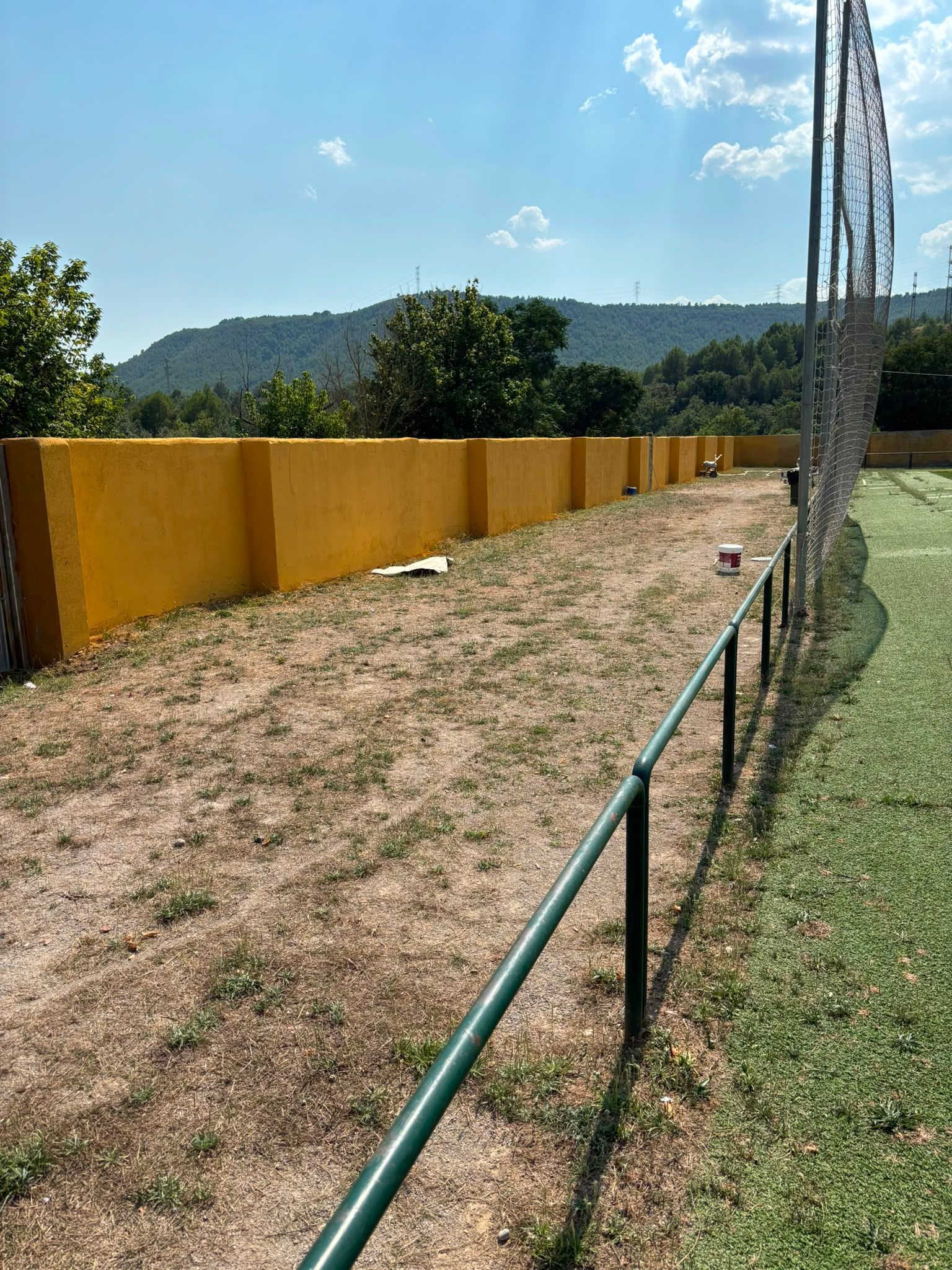 Sendero de tierra con paredes amarillas junto a una barandilla verde, con colinas y cielo azul al fondo.