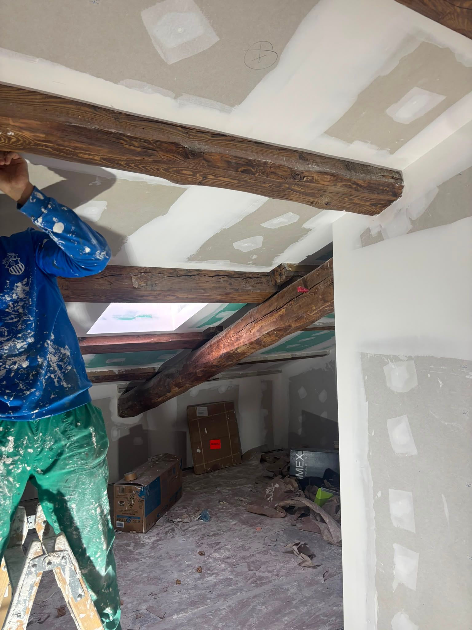 Un trabajador con sudadera azul instala una viga de madera en una buhardilla sin terminar.