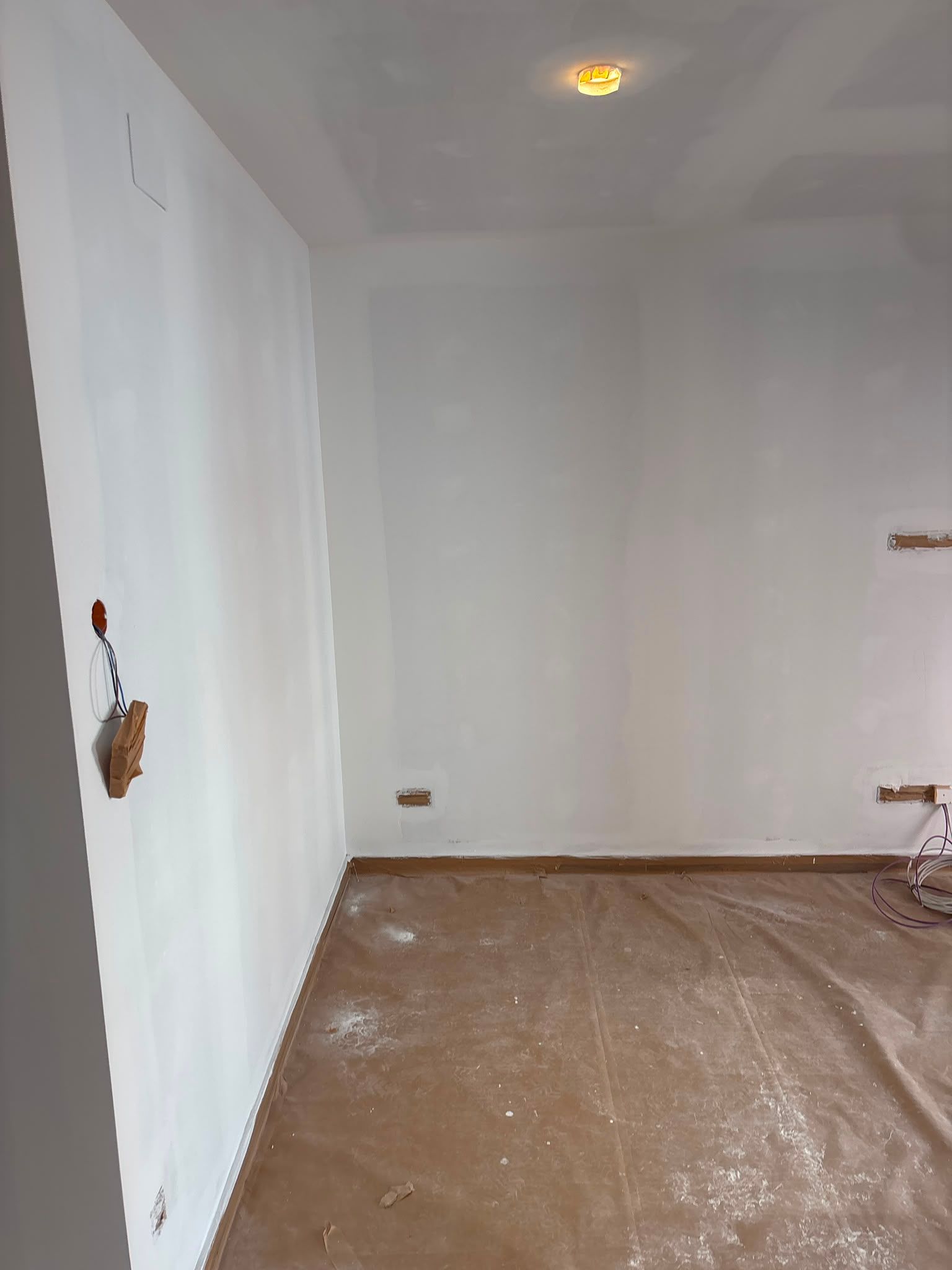 Habitación blanca vacía con suelo marrón sin terminar y una luz de techo encendida.