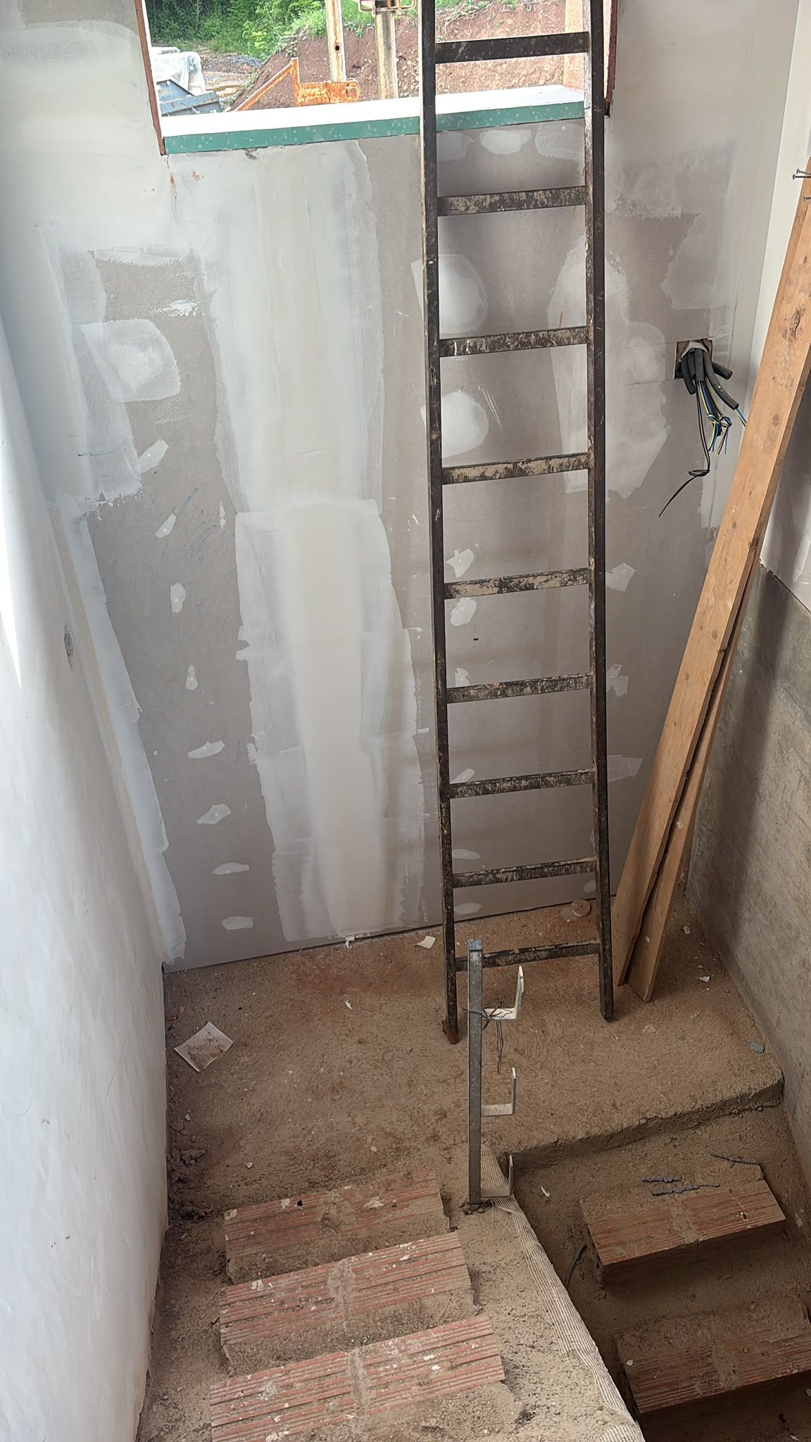Estrecha escalera sin terminar con paredes a la vista, una escalera metálica vertical y una pequeña ventana