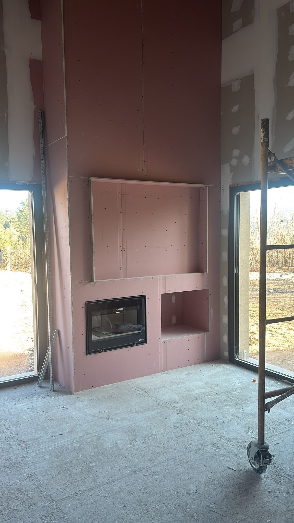 Nicho de chimenea sin terminar en una habitación con paredes de pladur a la vista y suelo de grava, junto a ventanas altas.