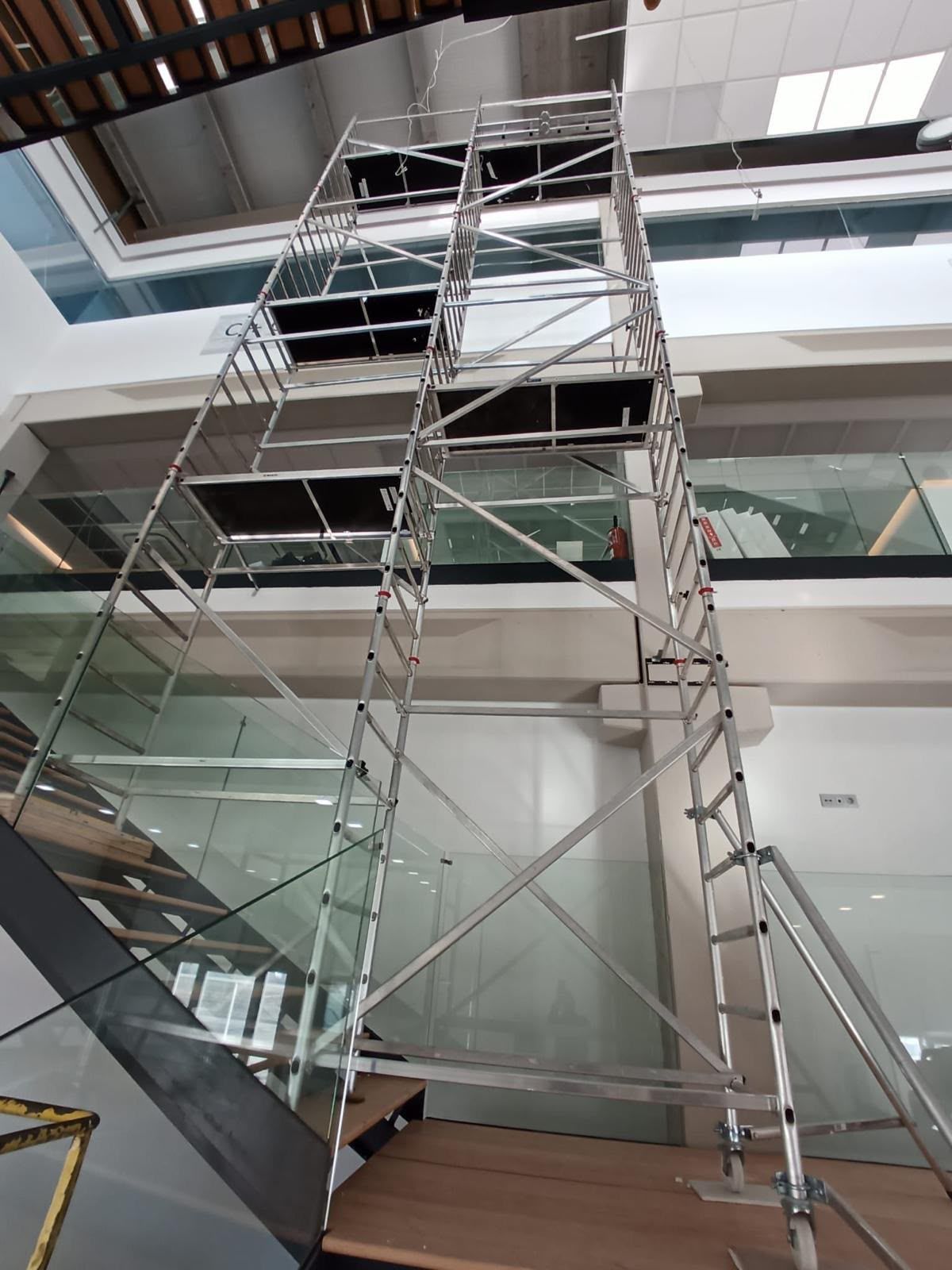 Andamios en el atrio de un edificio moderno, junto a una escalera y barandillas de cristal.
