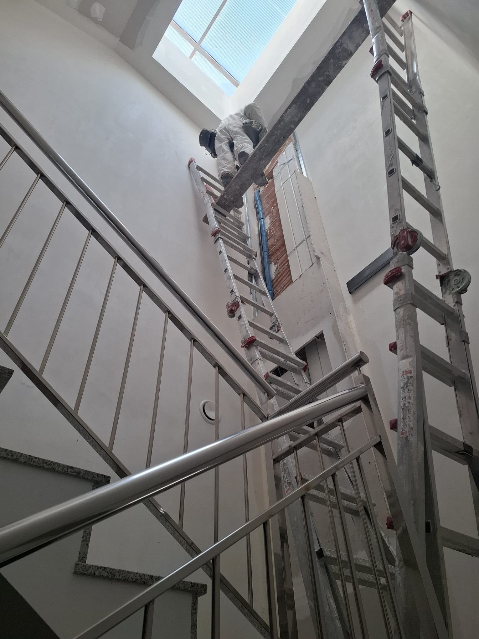 Persona subida a una escalera en un hueco de escalera, trabajando cerca del techo con otra escalera a su lado.
