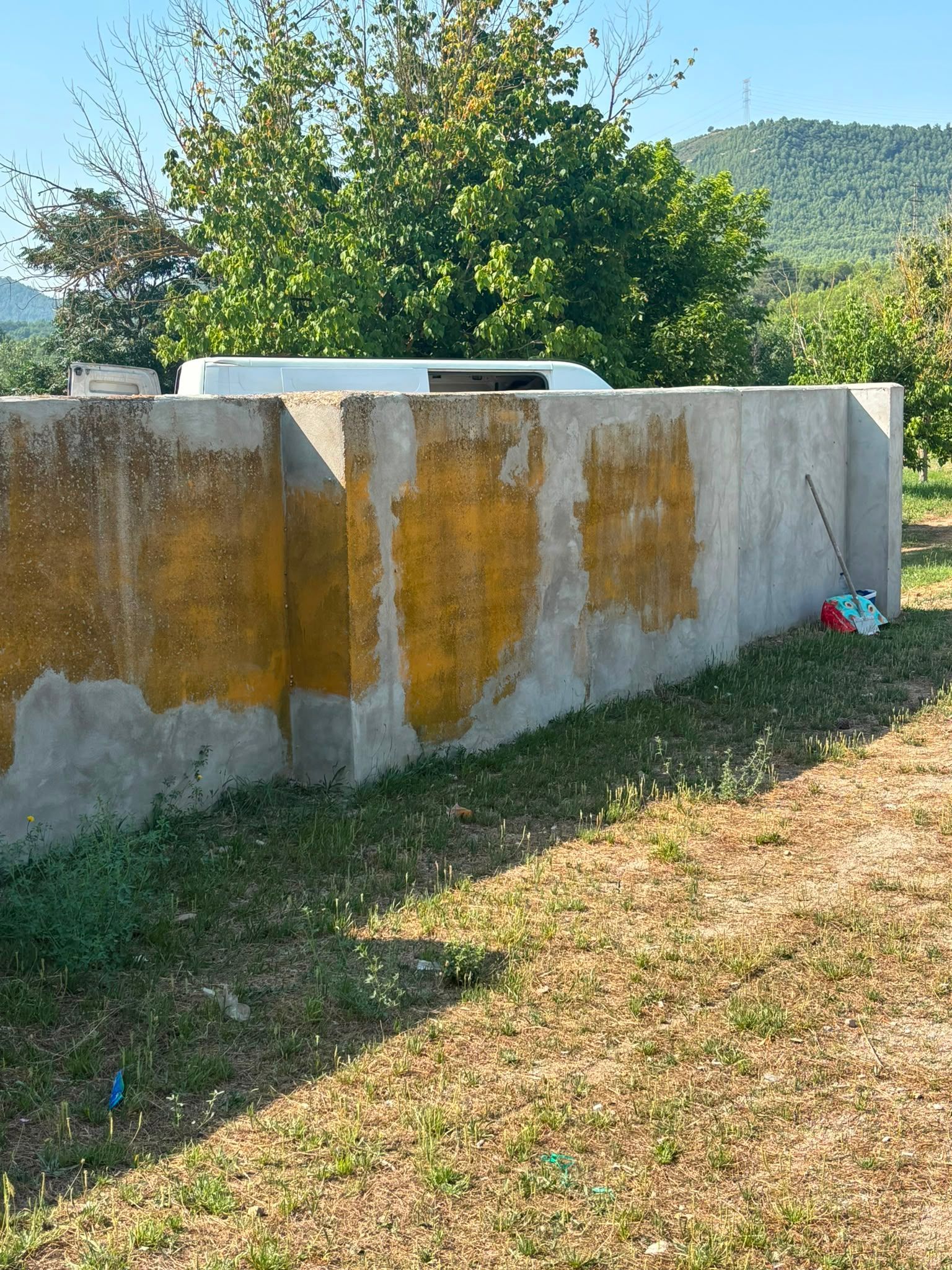Muro de hormigón con manchas amarillas junto a un jardín con césped, con árboles y colinas al fondo.