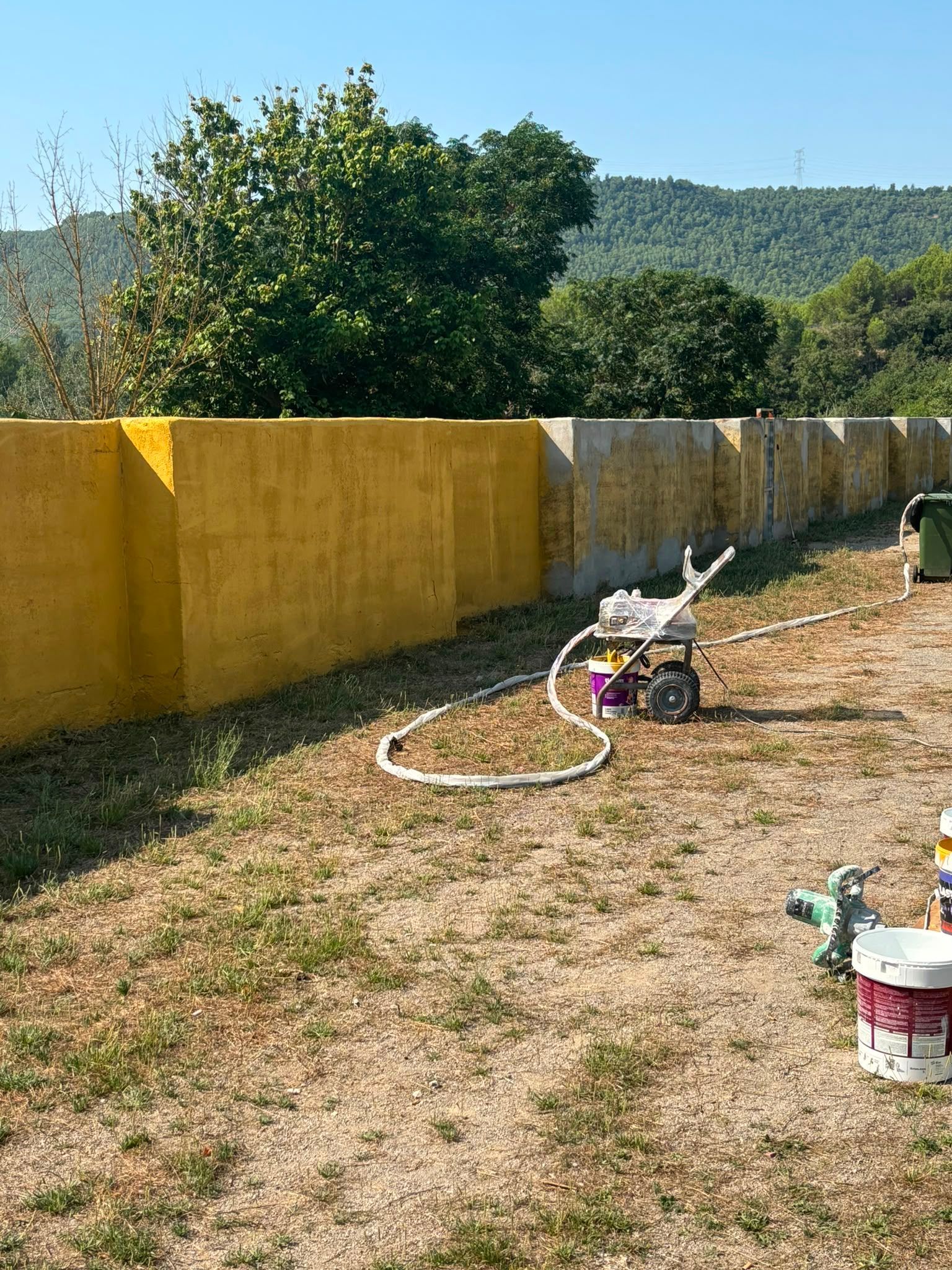Un muro amarillo bordea un patio seco con herramientas y cubos de pintura esparcidos frente a colinas boscosas.