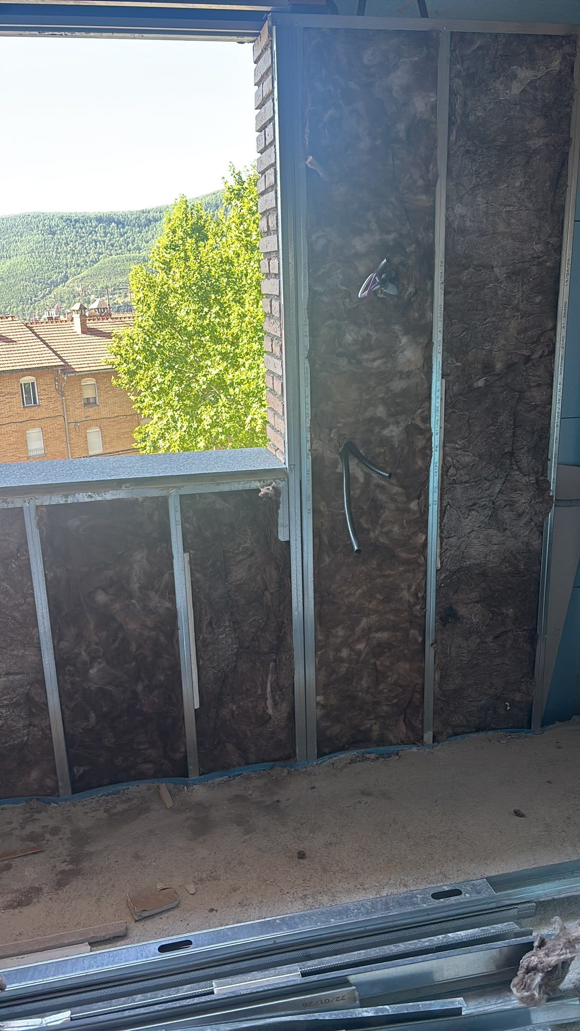 Pared de la habitación sin terminar con una ventana abierta que deja ver tejados y un árbol frondoso en el exterior.