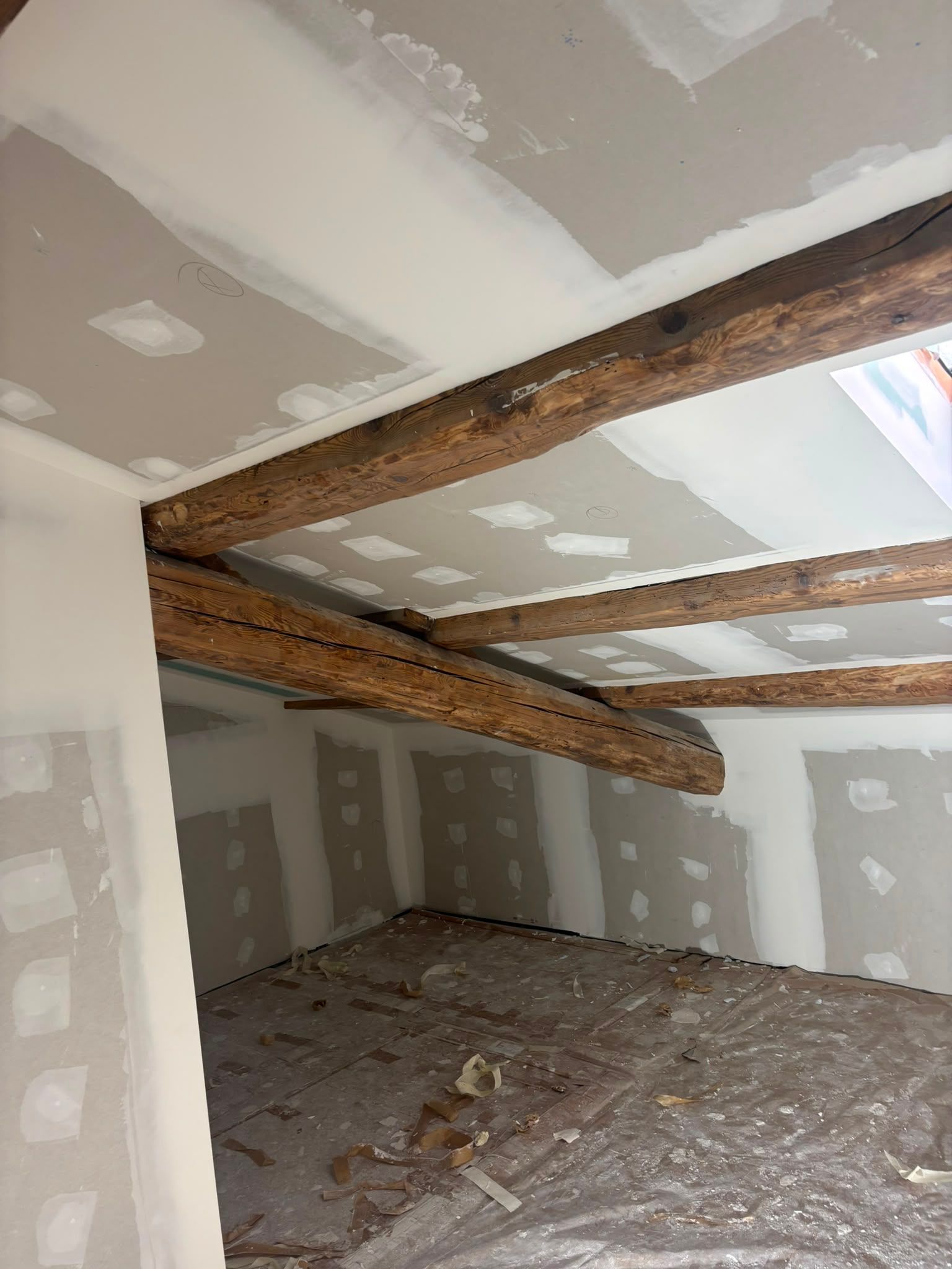 Habitación abuhardillada sin terminar, con paredes de pladur, vigas de madera a la vista y escombros en el suelo.