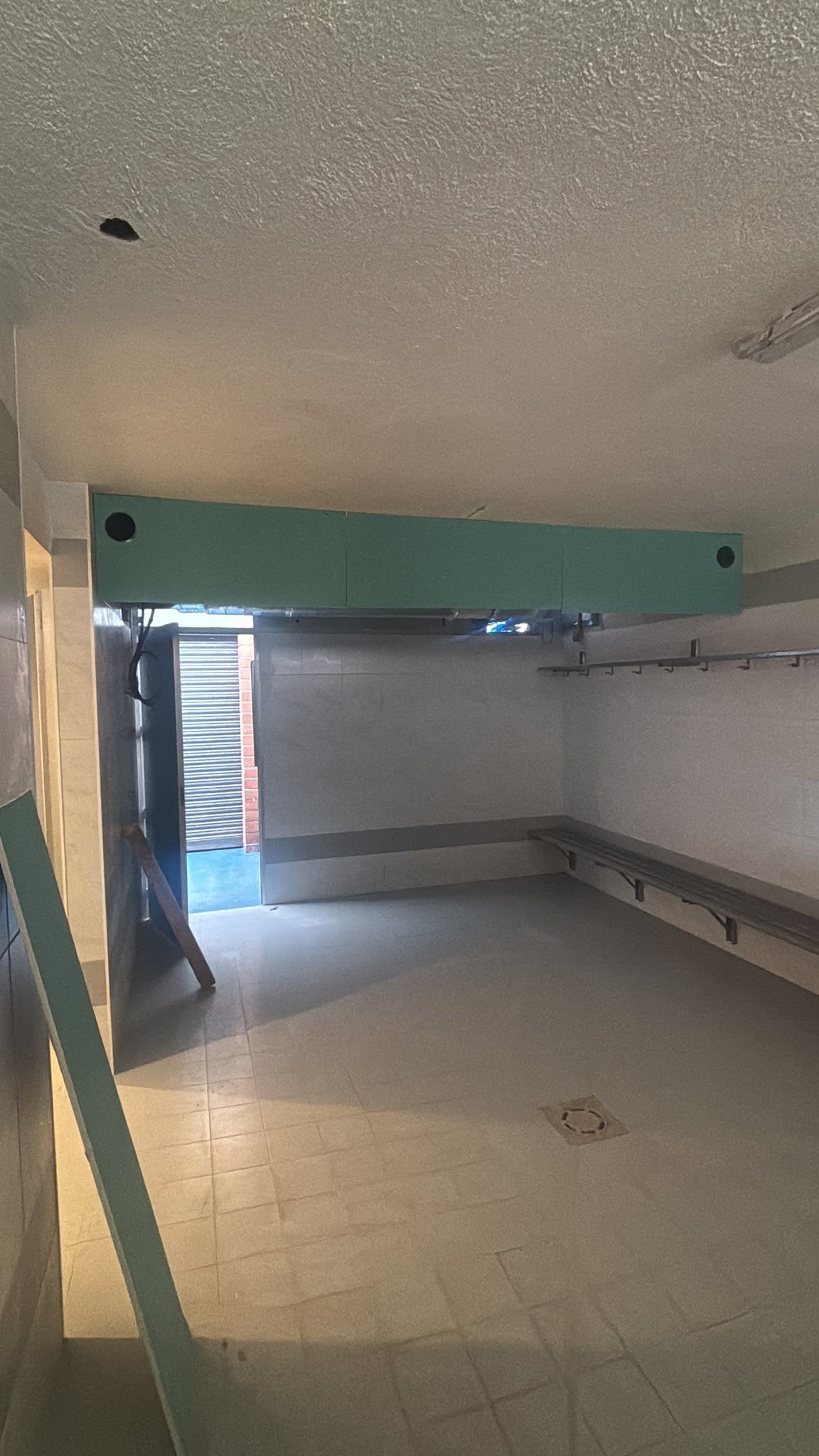 Habitación vacía con suelo de baldosas, techo bajo, viga de soporte verde y puerta abierta que da a las escaleras.