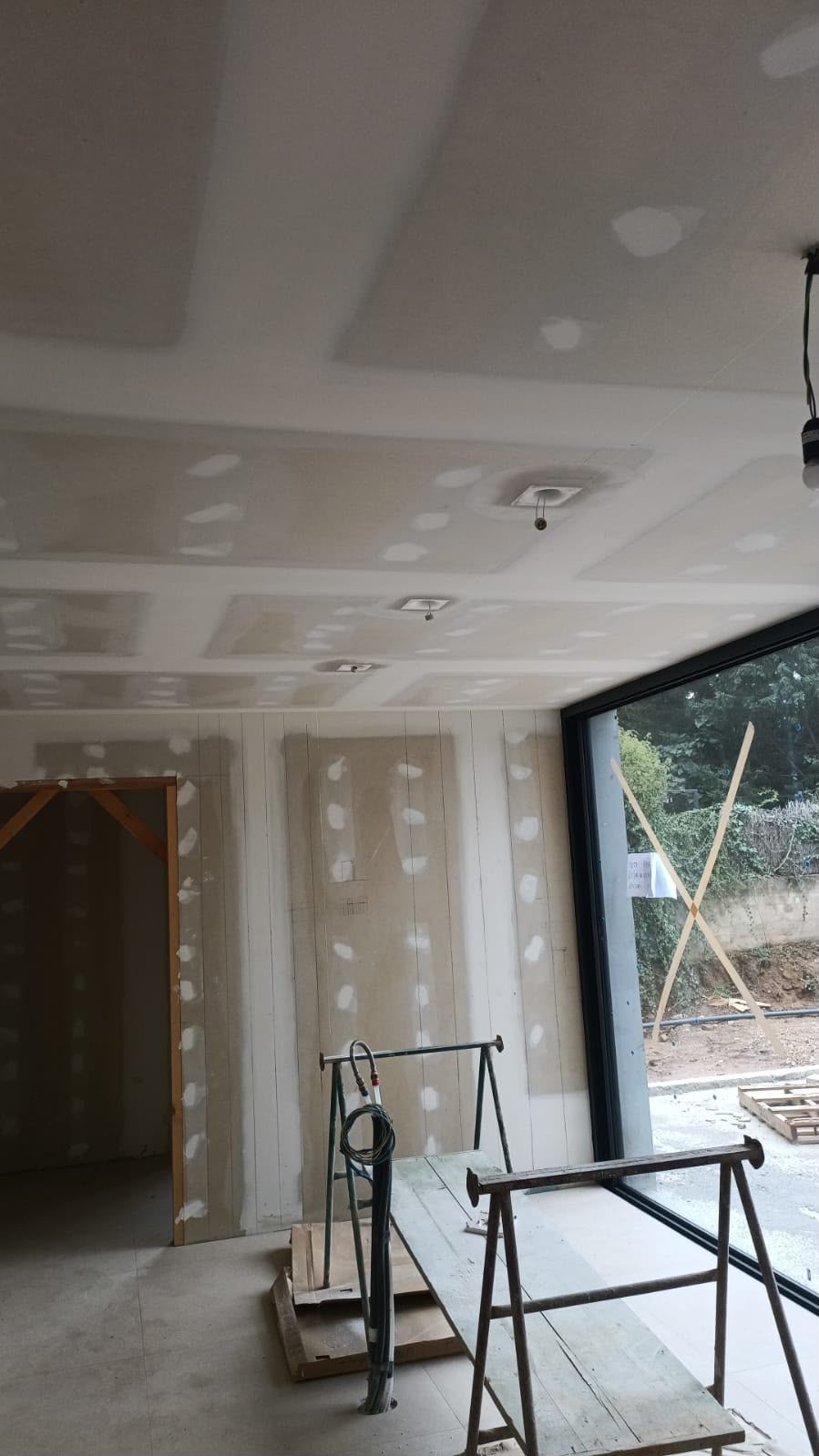 Habitación sin terminar con paneles de yeso, parches en el techo y dos mesitas plegables cerca de una gran ventana abierta.