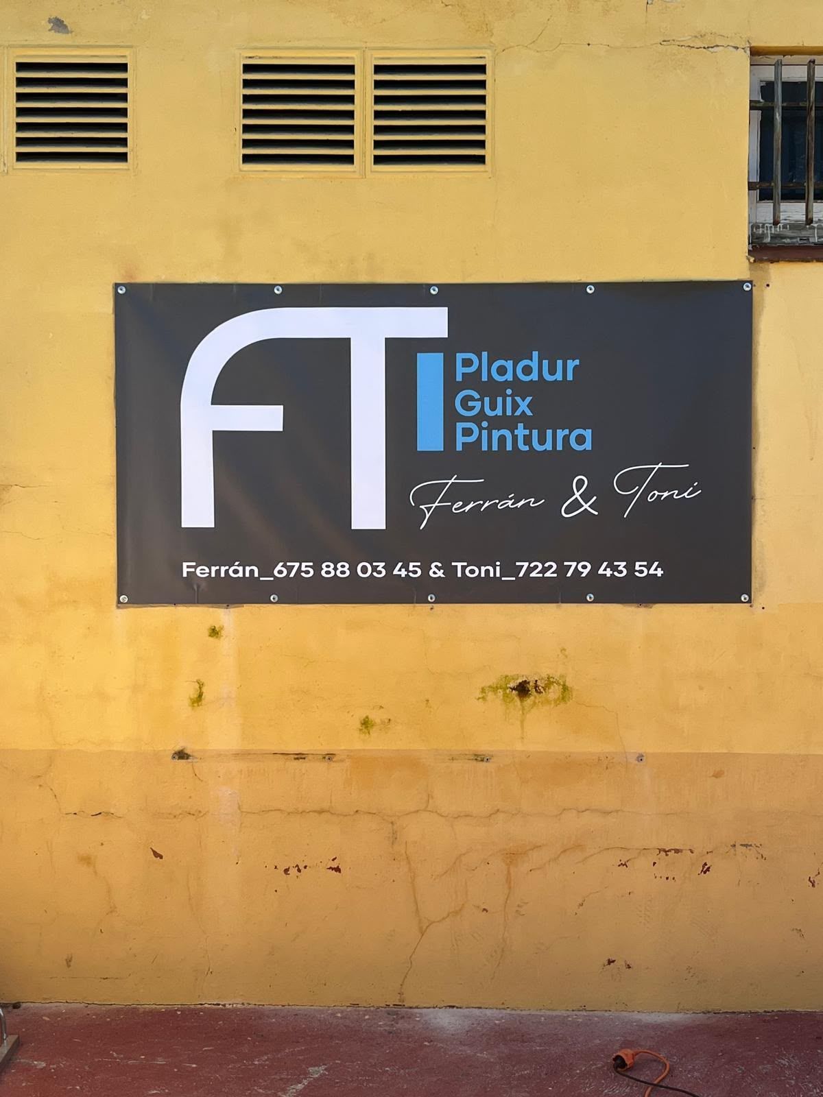 Letrero negro de “FT Pladur Gips Pintura” sobre una pared amarilla con rejillas de ventilación.