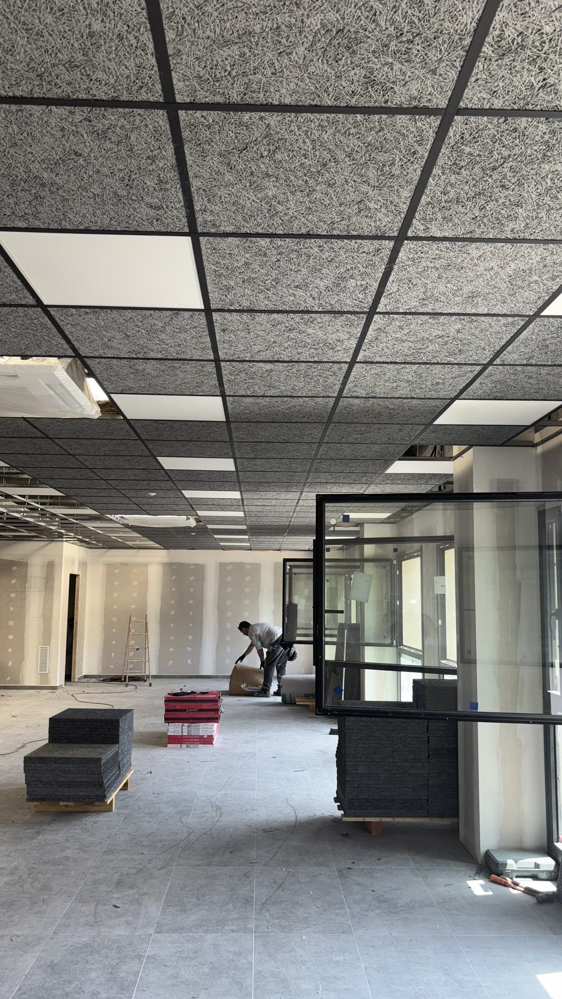 Oficina vacía con techo de baldosas a la vista, muebles dispersos y una mampara de cristal en primer plano.