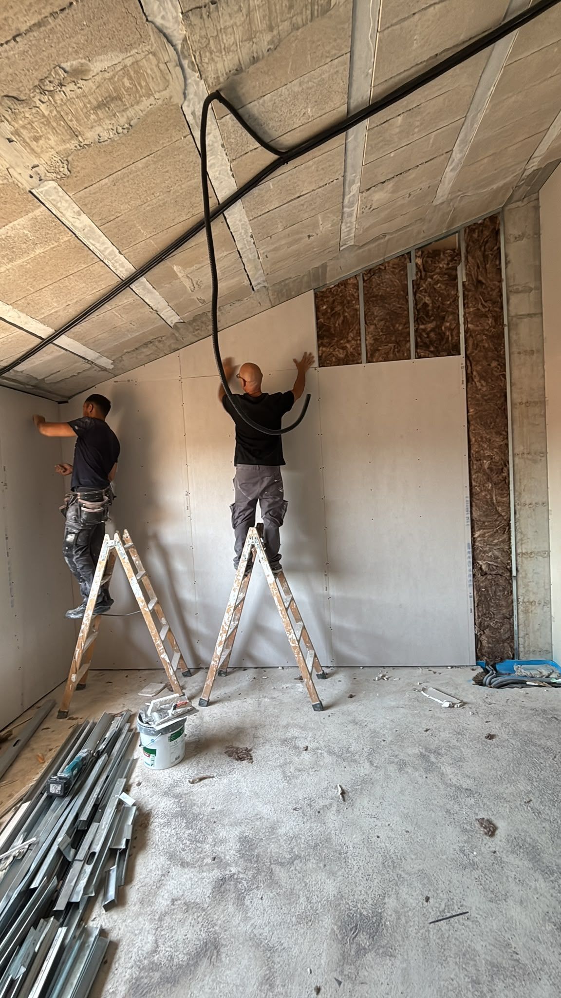 Dos trabajadores subidos a escaleras instalan paneles de yeso en el techo de una habitación parcialmente terminada.