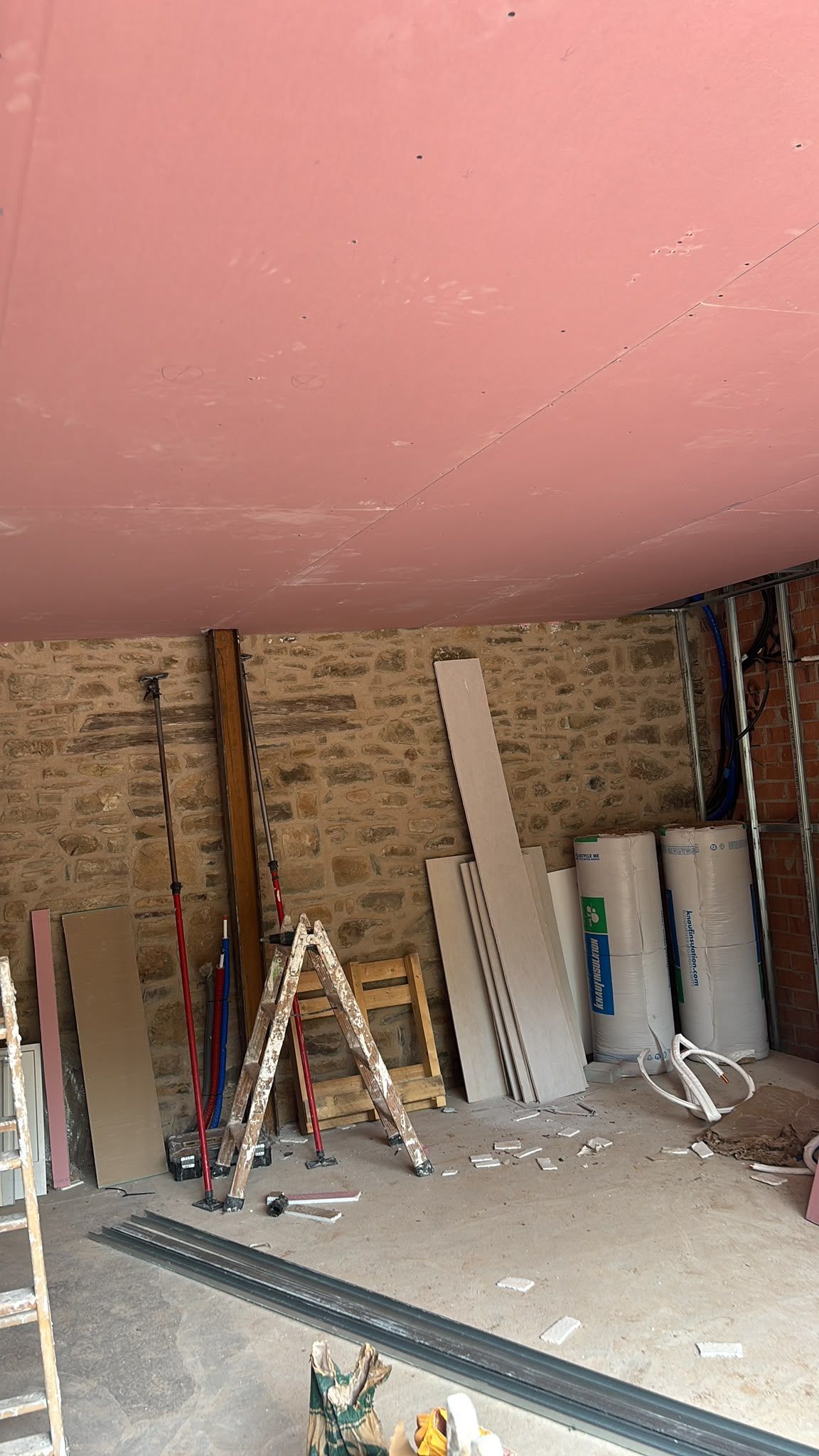 Habitación sin terminar con techo rosa, paredes de ladrillo visto, escaleras, tablones de madera