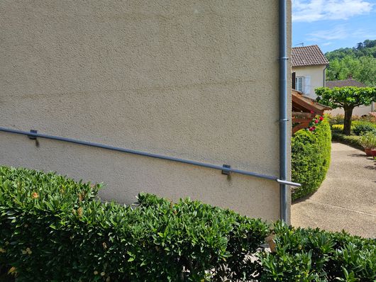 Côté d'une maison en stuc avec un tuyau métallique, une haie devant et une allée menant à un bâtiment carrelé.