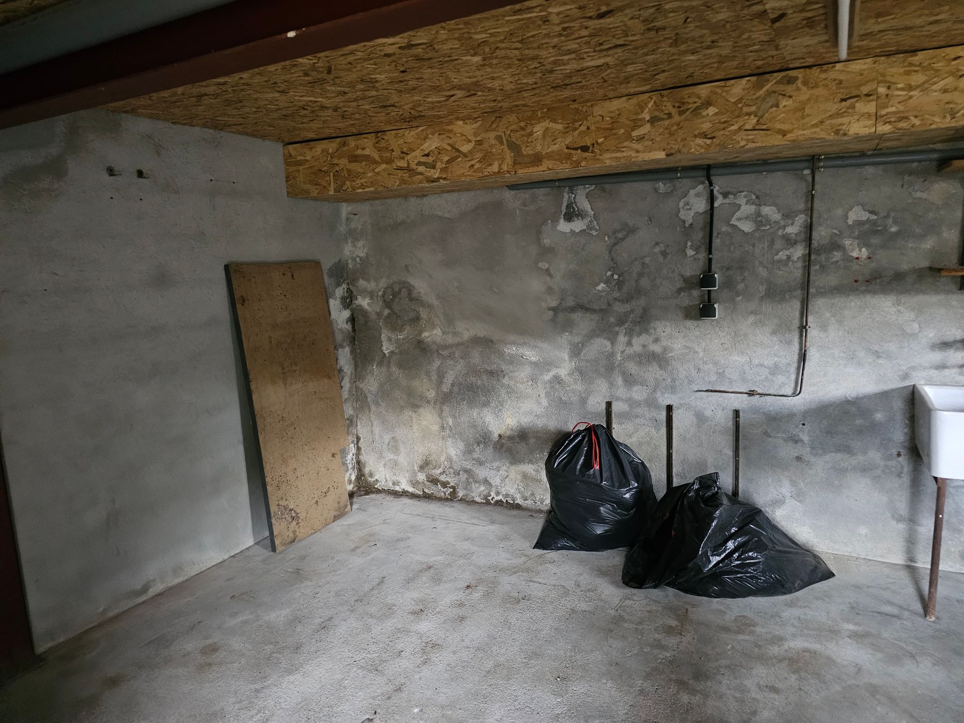 Sous-sol en béton avec murs qui s'effritent, panneaux de contreplaqué, sacs-poubelle noirs et poutres apparentes au plafond.