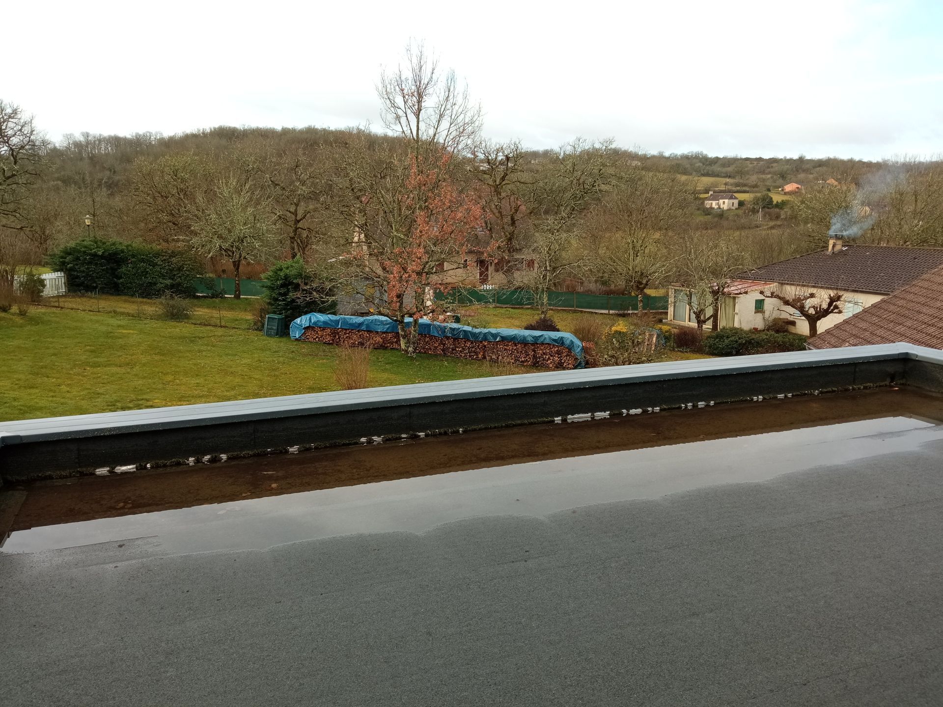 Vue depuis un toit sur un trottoir mouillé donnant sur une cour gazonnée, un tas de bois recouvert d'une bâche bleue et des arbres dénudés sous un ciel couvert.