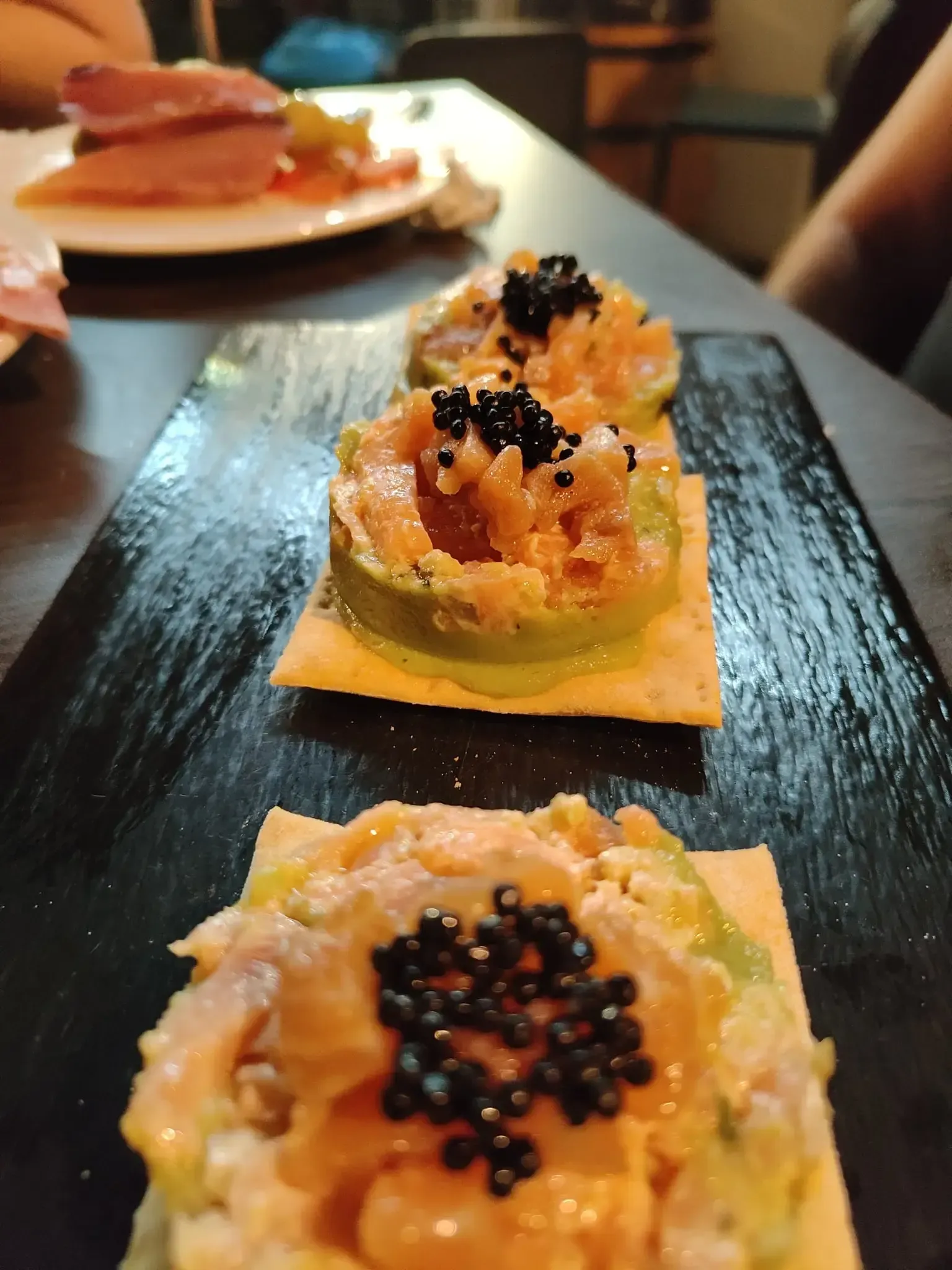 Aperitivo de sushi con huevas de naranja sobre aguacate, servido en un plato rectangular negro en un restaurante.