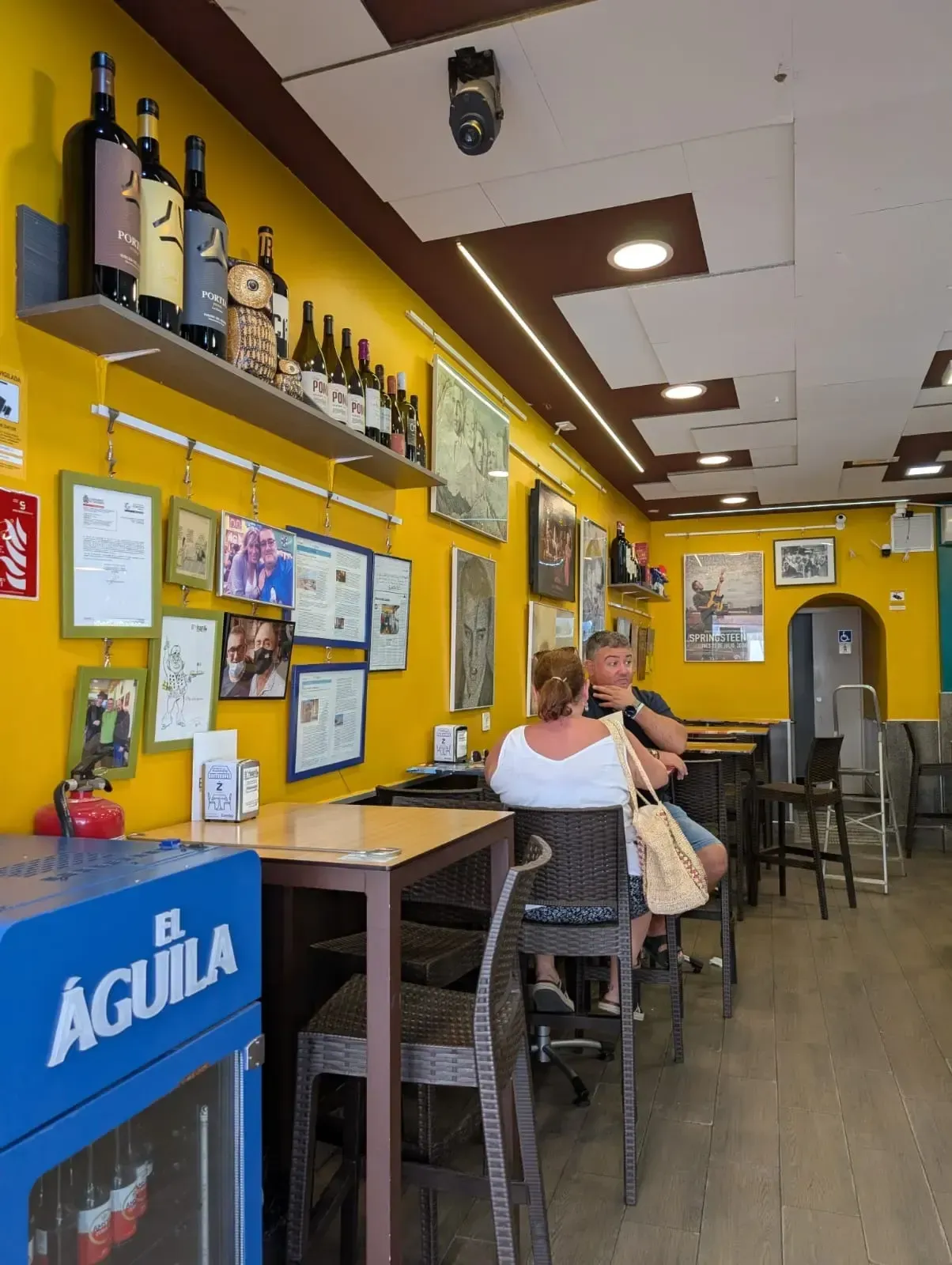 Interior de bar de color amarillo brillante con clientes sentados en mesas y botellas expuestas en estantes de la pared.