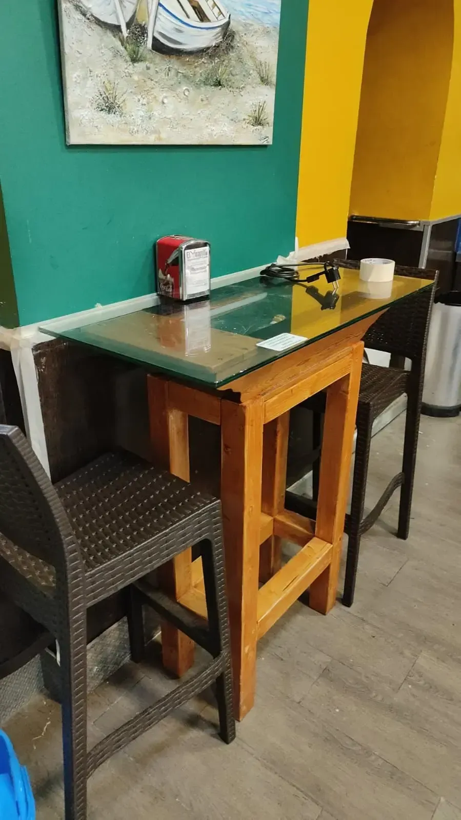 Una pequeña mesa de café con taburetes de madera, condimentos y bebidas, junto a una pared de color verde azulado y amarillo.