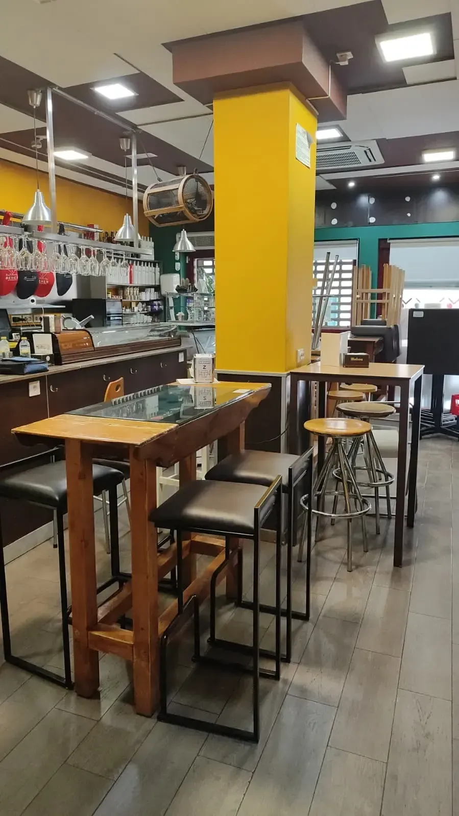 Interior vacío de una cafetería con mesas de madera, taburetes altos y una columna de soporte de color amarillo brillante.