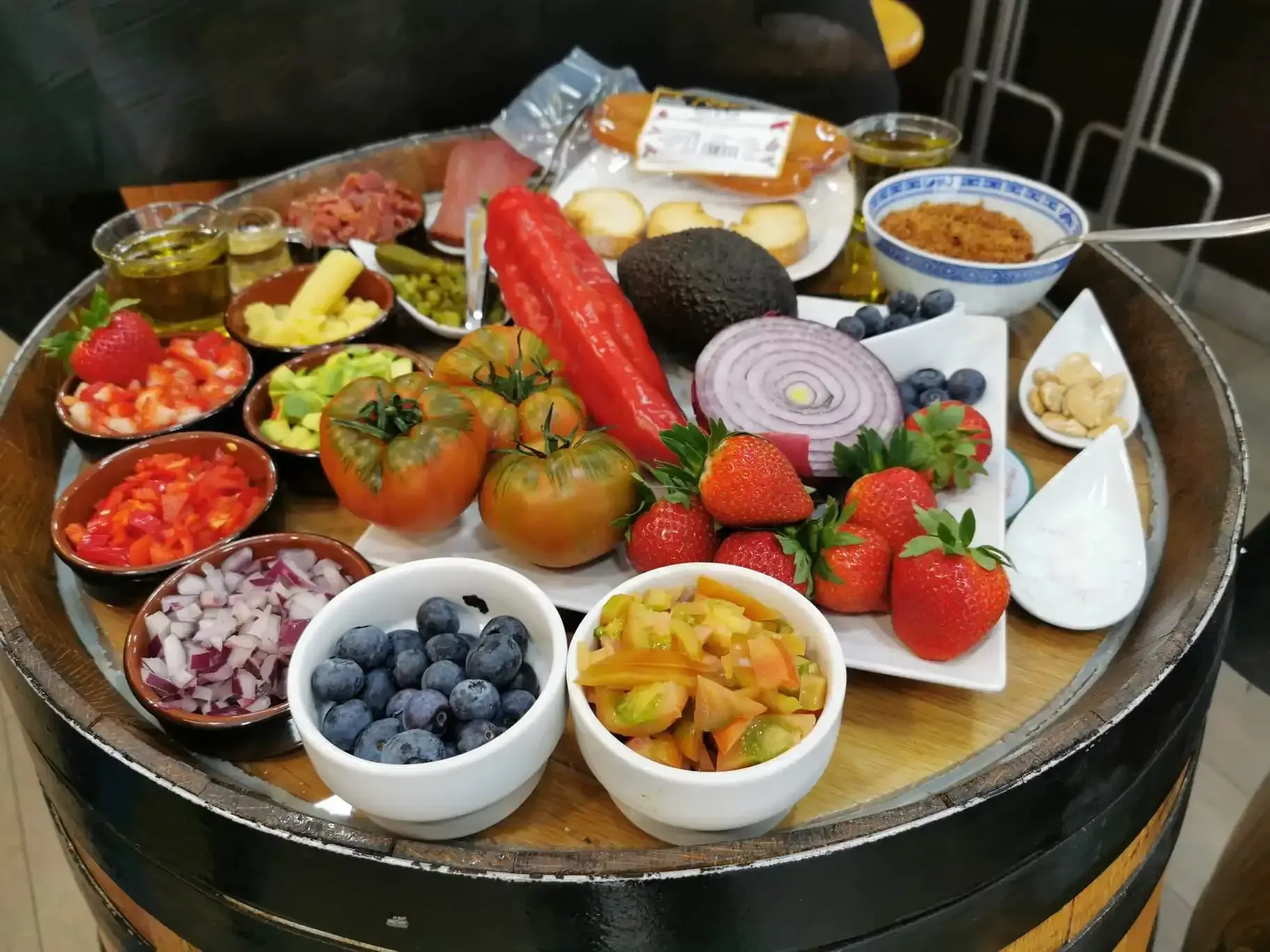 Variedad de frutas frescas, verduras, quesos y salsas dispuestas sobre una mesa redonda al aire libre.
