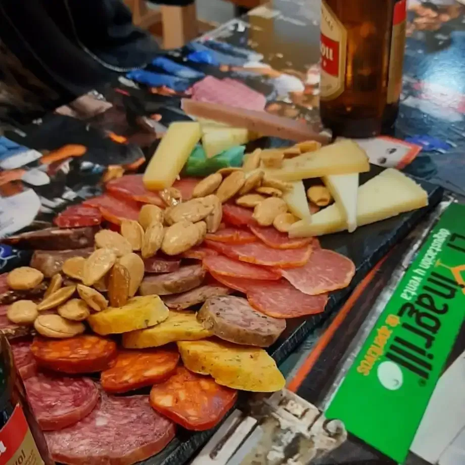 Tabla de embutidos con carnes en lonchas, galletas saladas, frutos secos, queso y bebidas sobre una mesa.