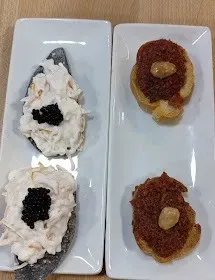 Aperitivo emplatado con cuatro pequeños ingredientes: dos bocados blancos y negros a la izquierda, dos bocados rojos