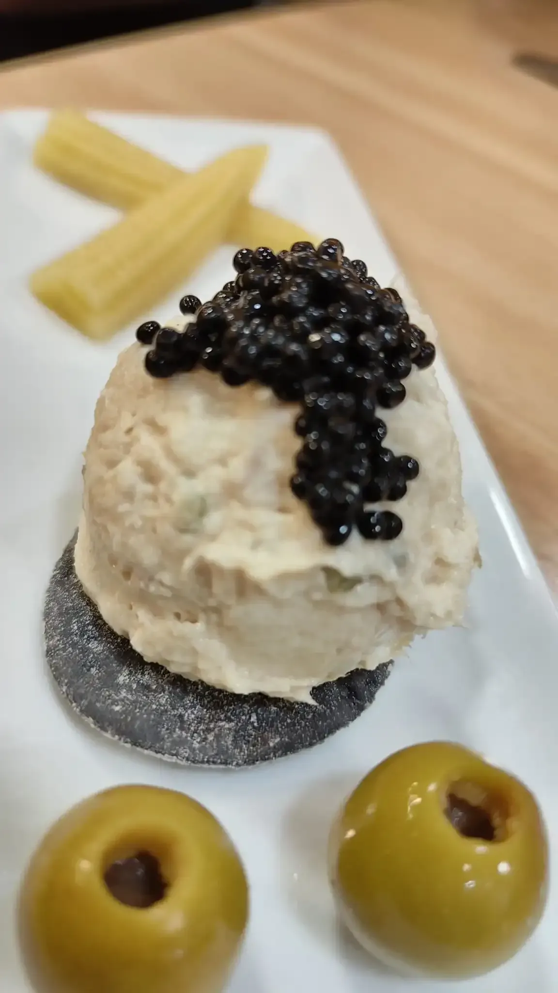 Postre blanco coronado con caviar negro sobre una base metálica, con aceitunas y trozos crujientes a su lado.