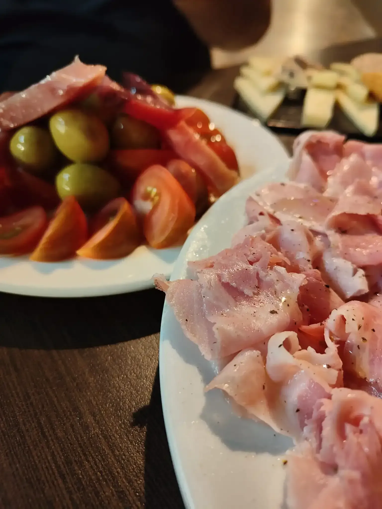 Platos de jamón en lonchas y ensalada de tomate y aceitunas sobre una mesa oscura.