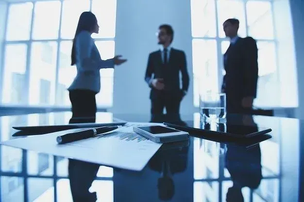 Reunión de negocios en una oficina moderna. Tres personas conversan, concentradas en documentos, teléfono y bolígrafos sobre la mesa.