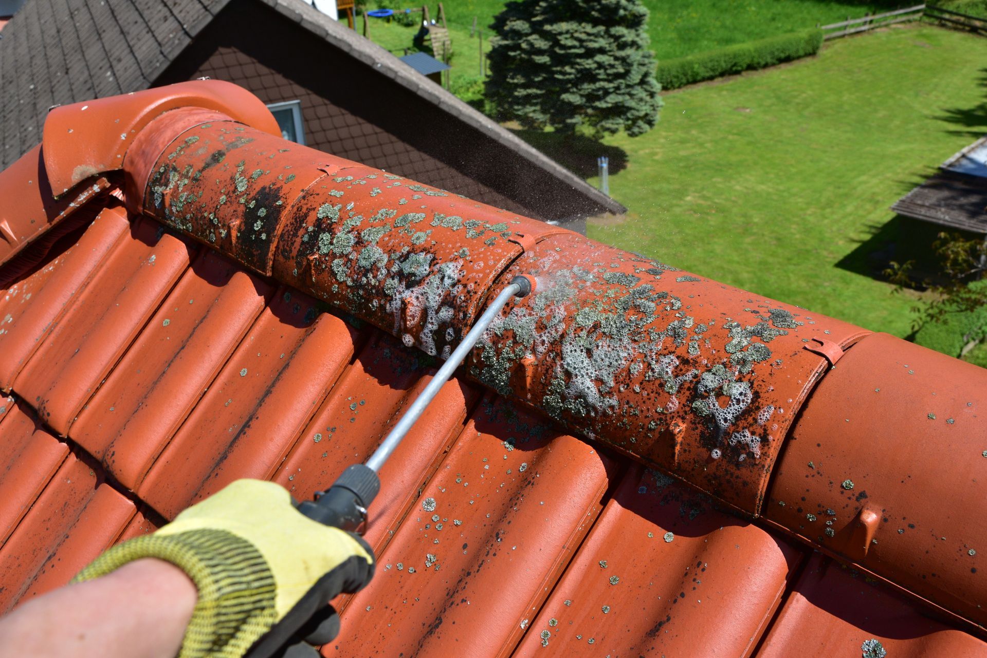 Une main gantée tenant un nettoyeur haute pression pour nettoyer le lichen d'une crête de tuiles en terre cuite rouge.