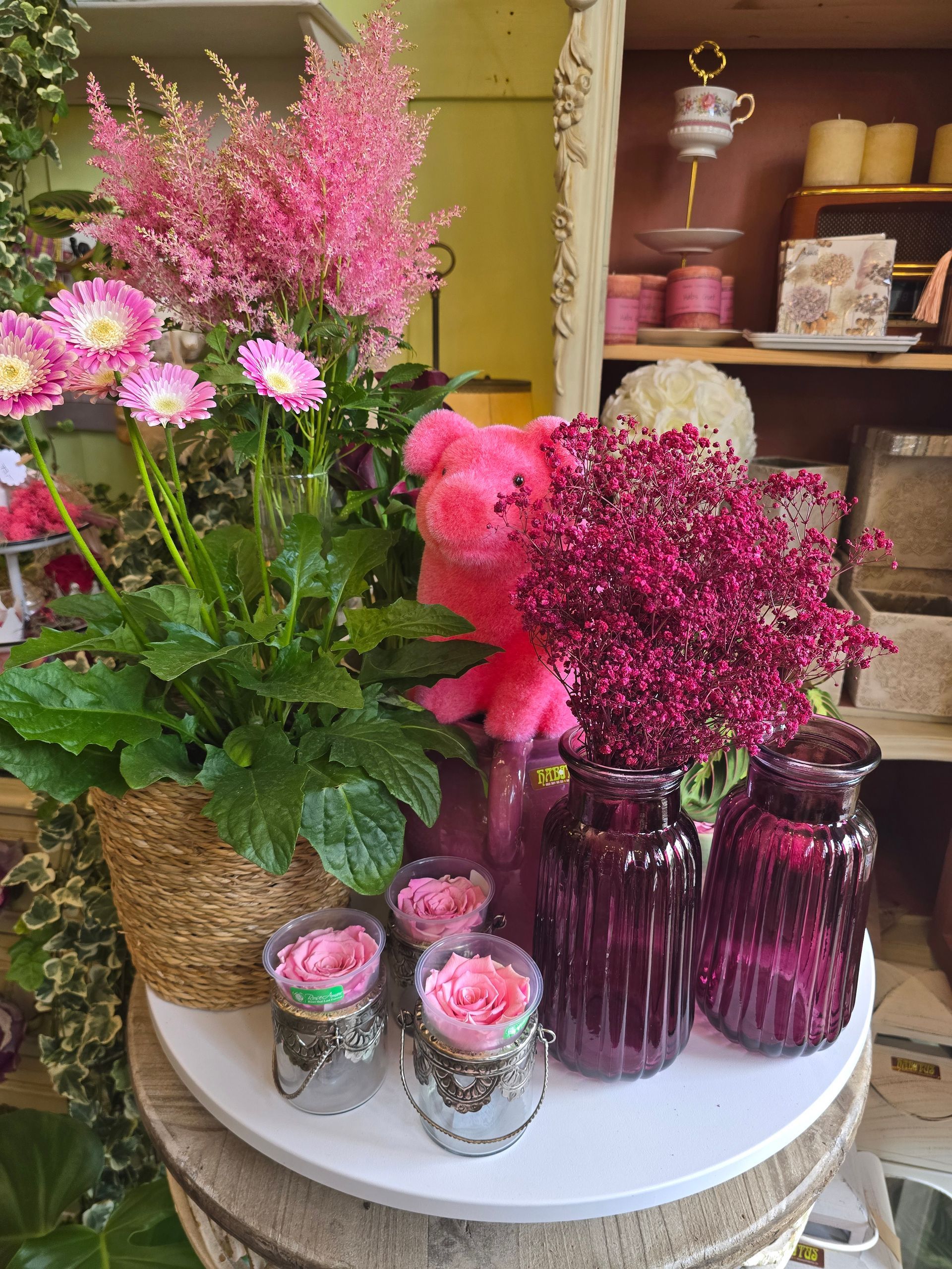 Ein rosa Teddybär sitzt neben einer Vase mit rosa Blumen auf einem Tisch.