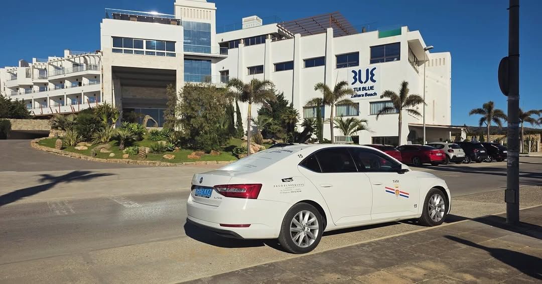 Sedán blanco en una calle frente a un edificio blanco de un complejo turístico con letreros azules.