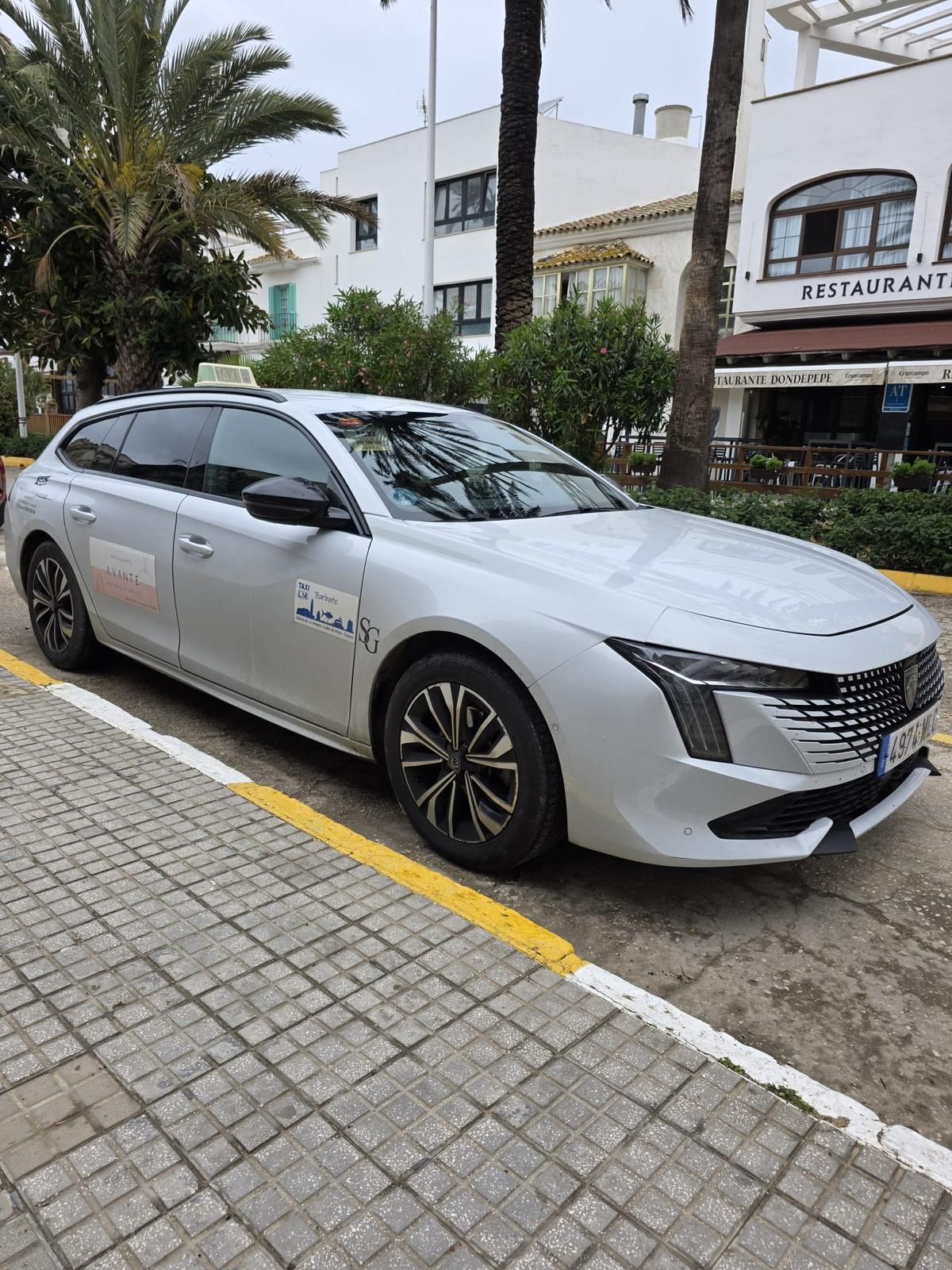 Un sedán blanco estacionado junto a la acera en una calle de la ciudad con edificios y palmeras cerca.