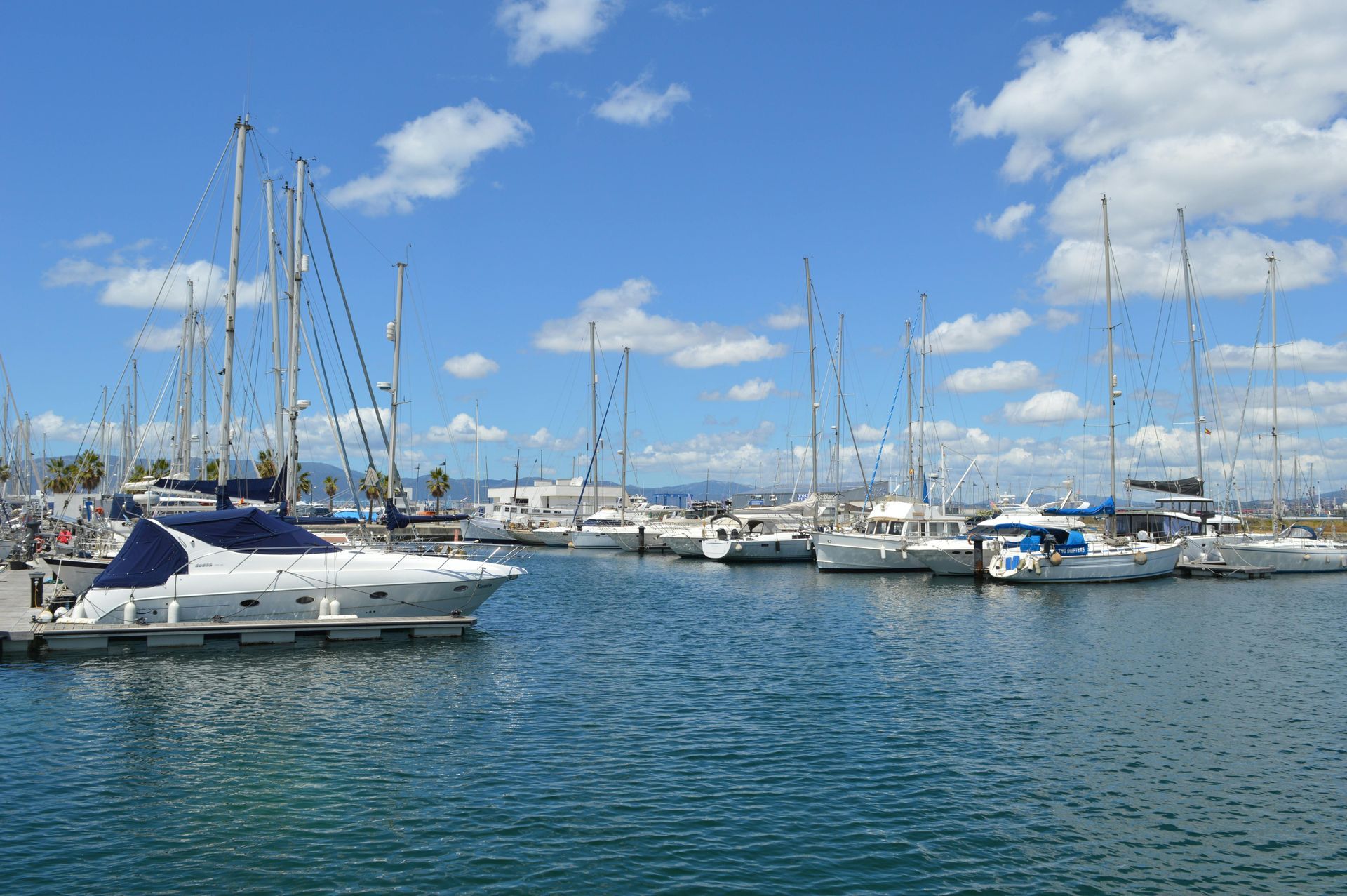 Veleros y yates amarrados en un puerto deportivo soleado bajo un cielo azul con nubes dispersas.