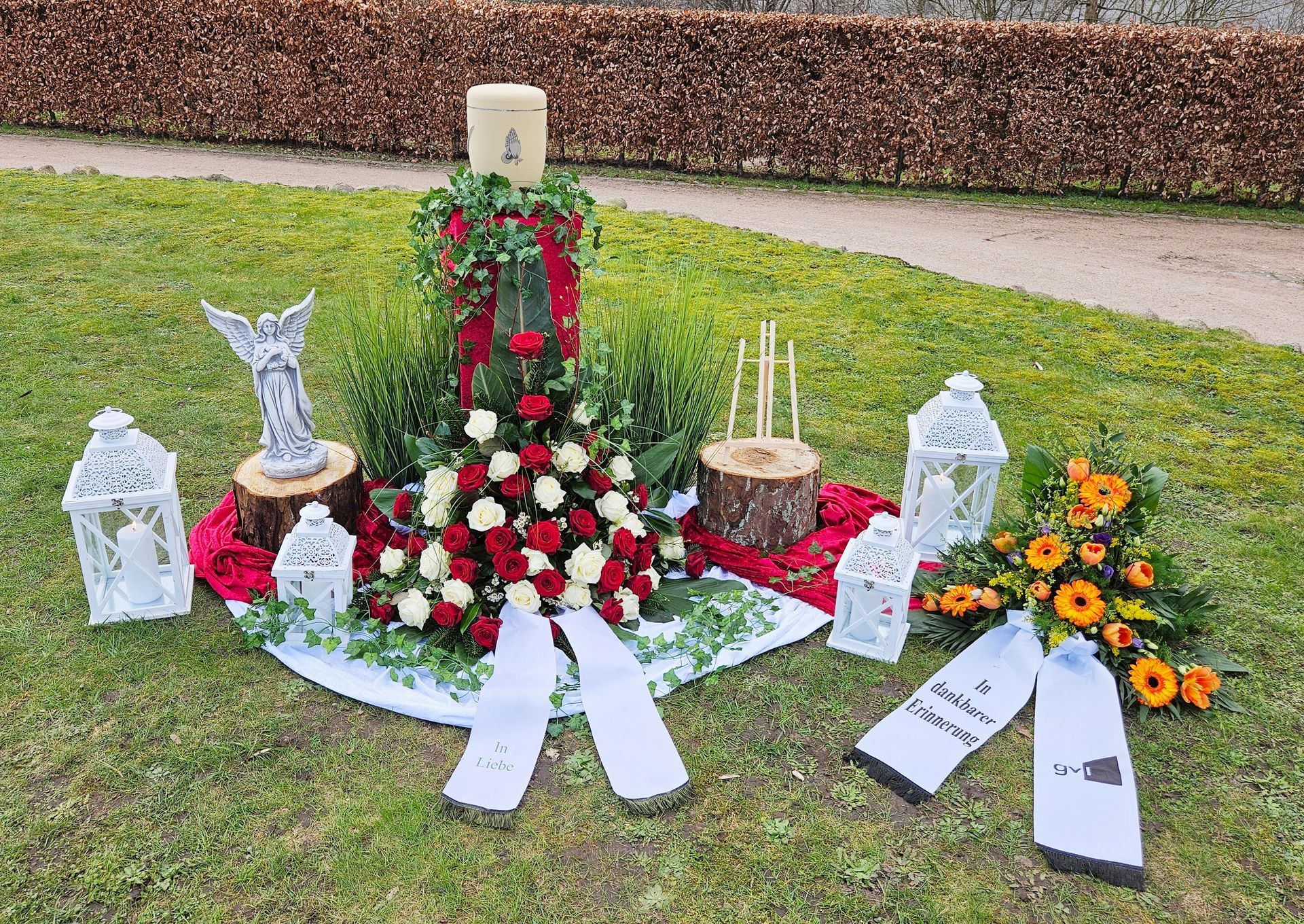 Ein Denkmal mit Blumen, Kerzen, Laternen und einer Statue im Gras.