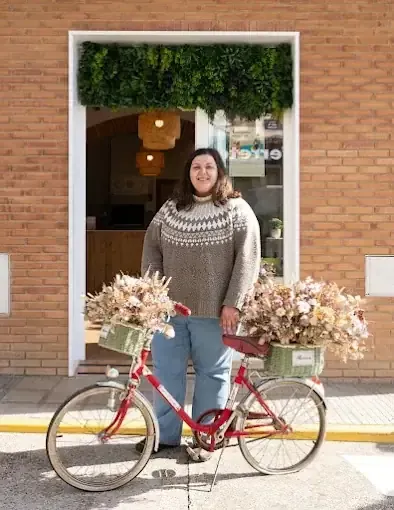 Una mujer se encuentra junto a una bicicleta roja con cestas de flores, frente a una tienda con follaje verde.
