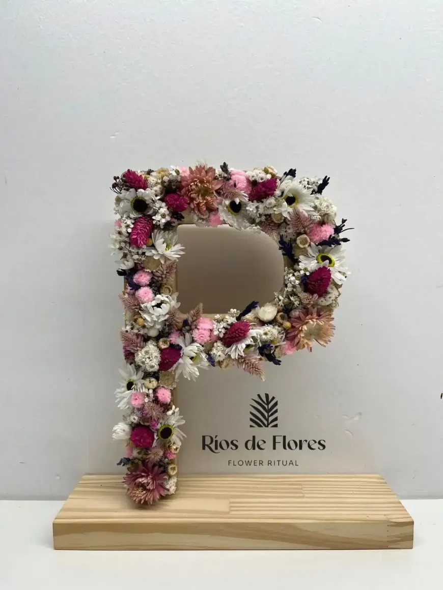 Arreglo floral en forma de letra “P” sobre soporte de madera, con flores de color rosa, blanco y burdeos.