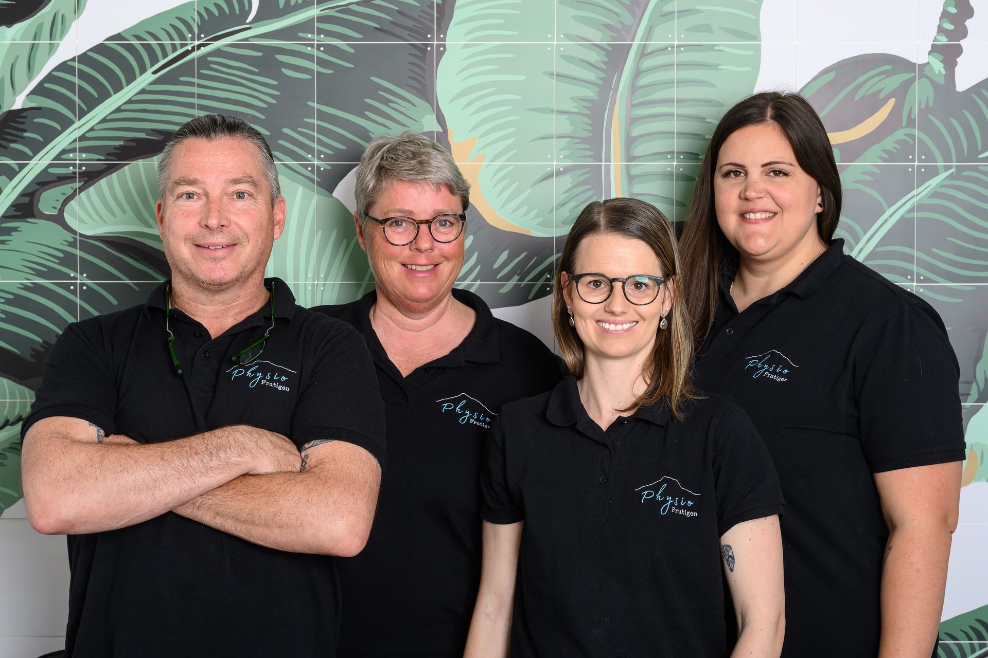 Vier lächelnde Teammitglieder in schwarzen Poloshirts stehen vor einem laubbedeckten Wandgemälde