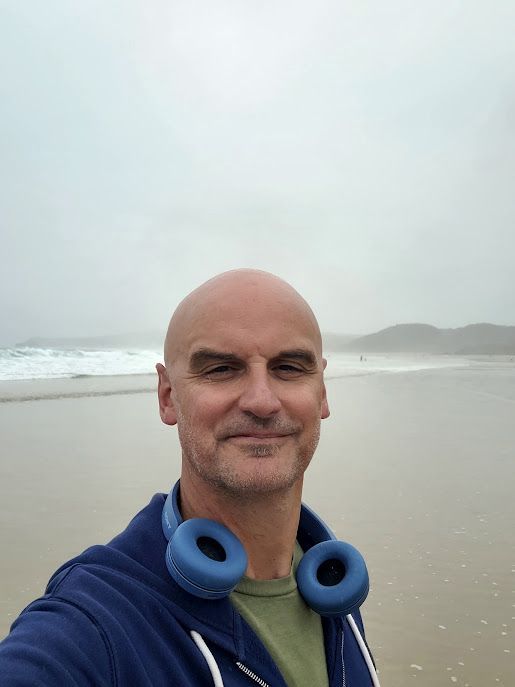 Hombre calvo en la playa, con auriculares azules al cuello, sonriendo. Cielo nublado y océano de fondo.