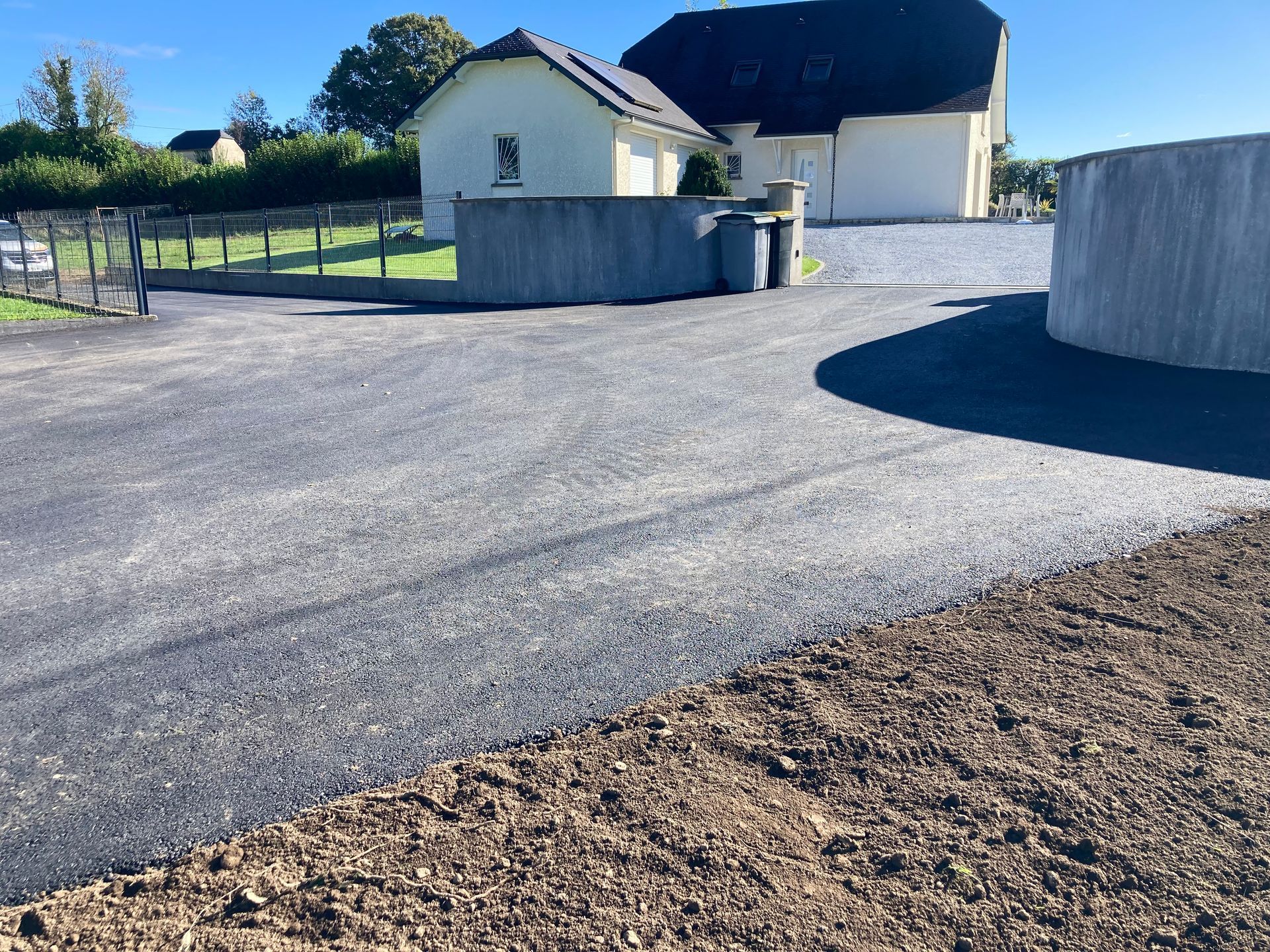 Allée asphaltée menant à une maison de couleur claire avec un toit foncé, entourée de pelouse et de clôtures.