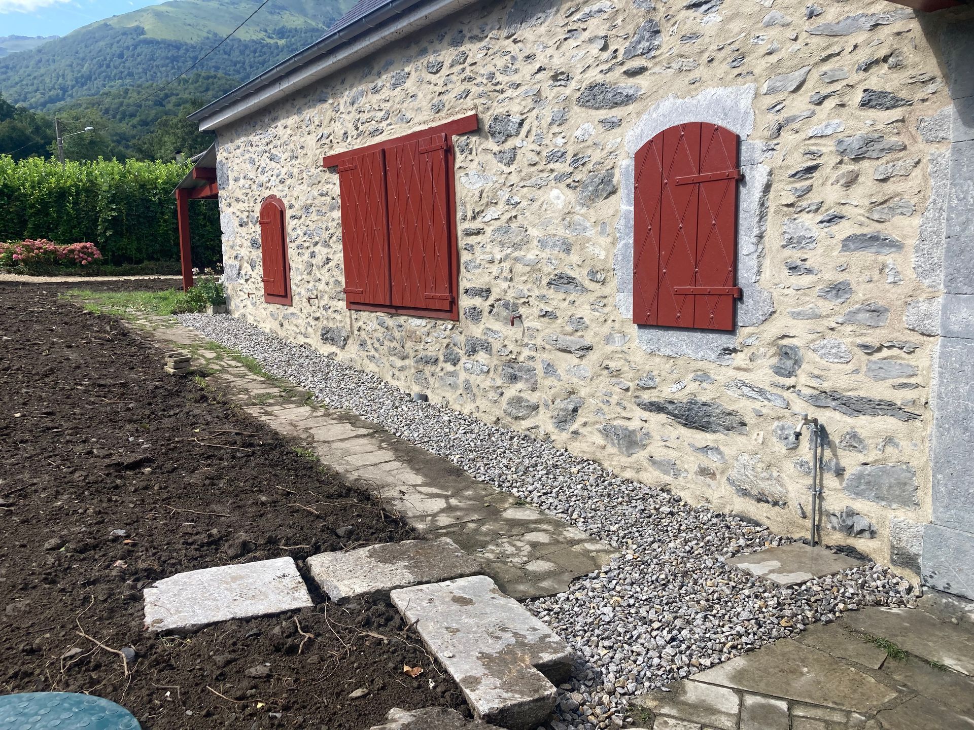 Bâtiment en pierre aux volets rouges ; chemin de gravier et jardin.