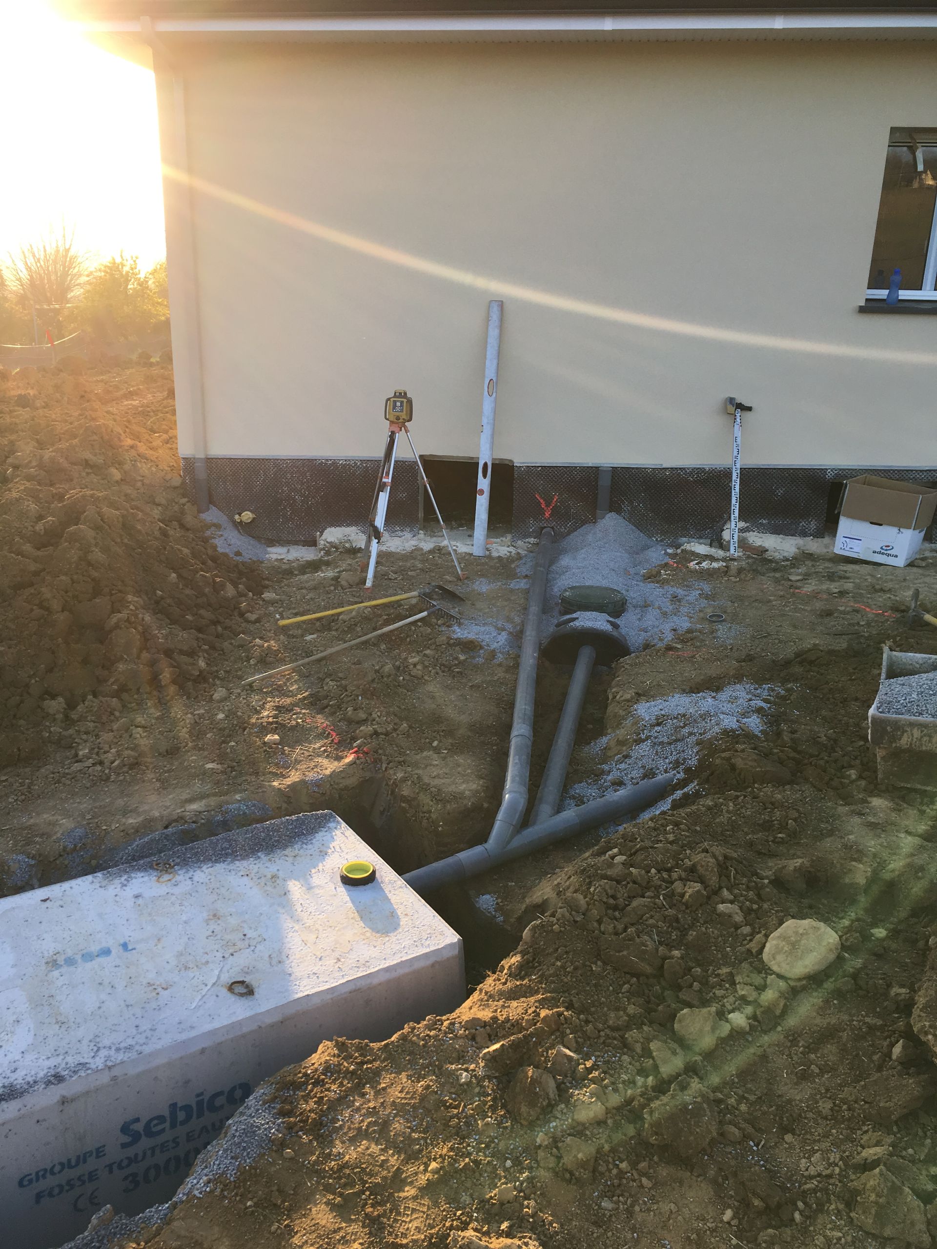 Chantier de construction avec une cuve en béton dans une tranchée près d'un bâtiment. Des tuyaux et un niveau sont visibles.