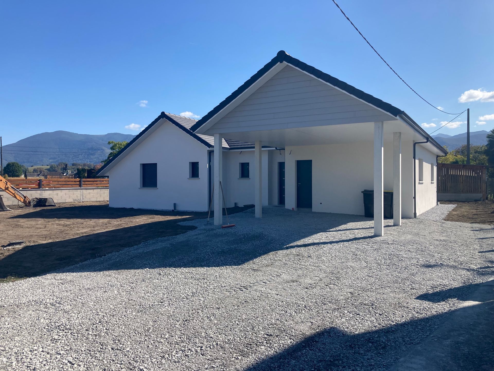 Maison blanche avec abri de voiture sur allée de gravier, montagnes en arrière-plan. Ciel bleu.