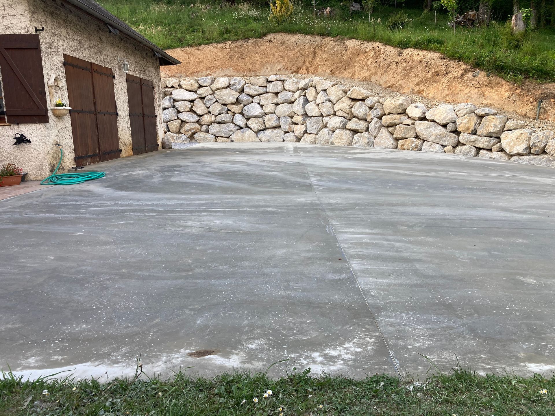 Terrasse en béton gris devant un mur de soutènement en pierre et une maison beige aux portes marron.