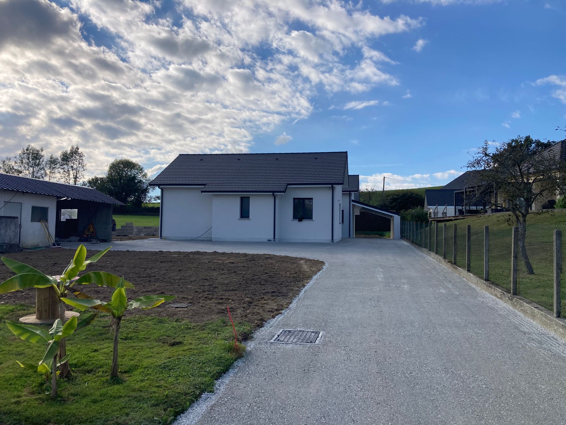 Maison blanche au toit sombre, allée de gravier, ciel bleu nuageux.