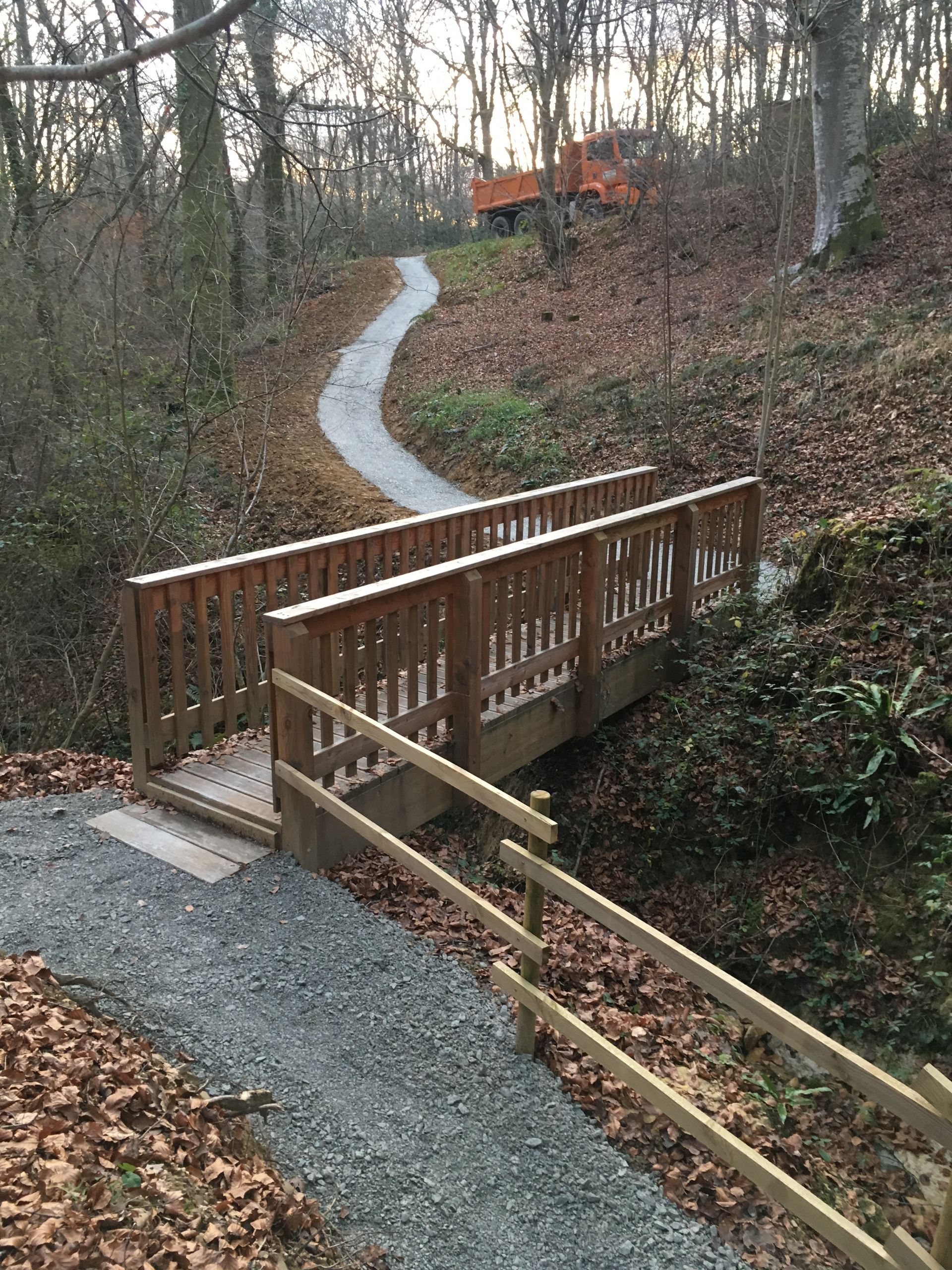 Pont de bois enjambant un petit ruisseau, menant à un chemin de gravier sinueux traversant une zone boisée.